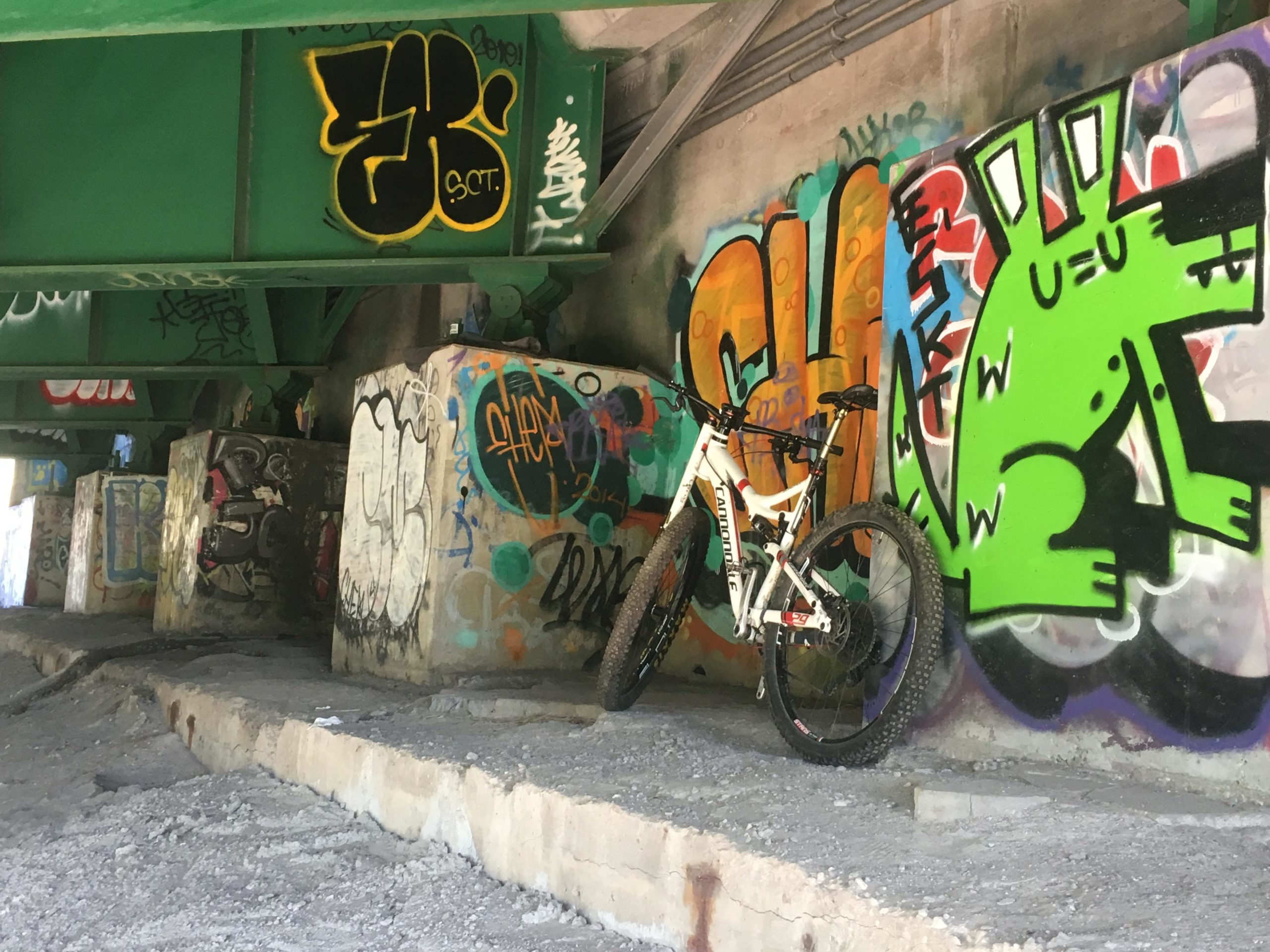 A white mountain bike is leaned against a wall covered in vibrant graffiti, including a large green cartoonish creature. The scene is set under a concrete overpass, with colorful tags and designs on the pillars and walls, showcasing urban art. The ground is uneven and strewn with gravel, highlighting the rugged environment. Don Valley mountain bike trail.