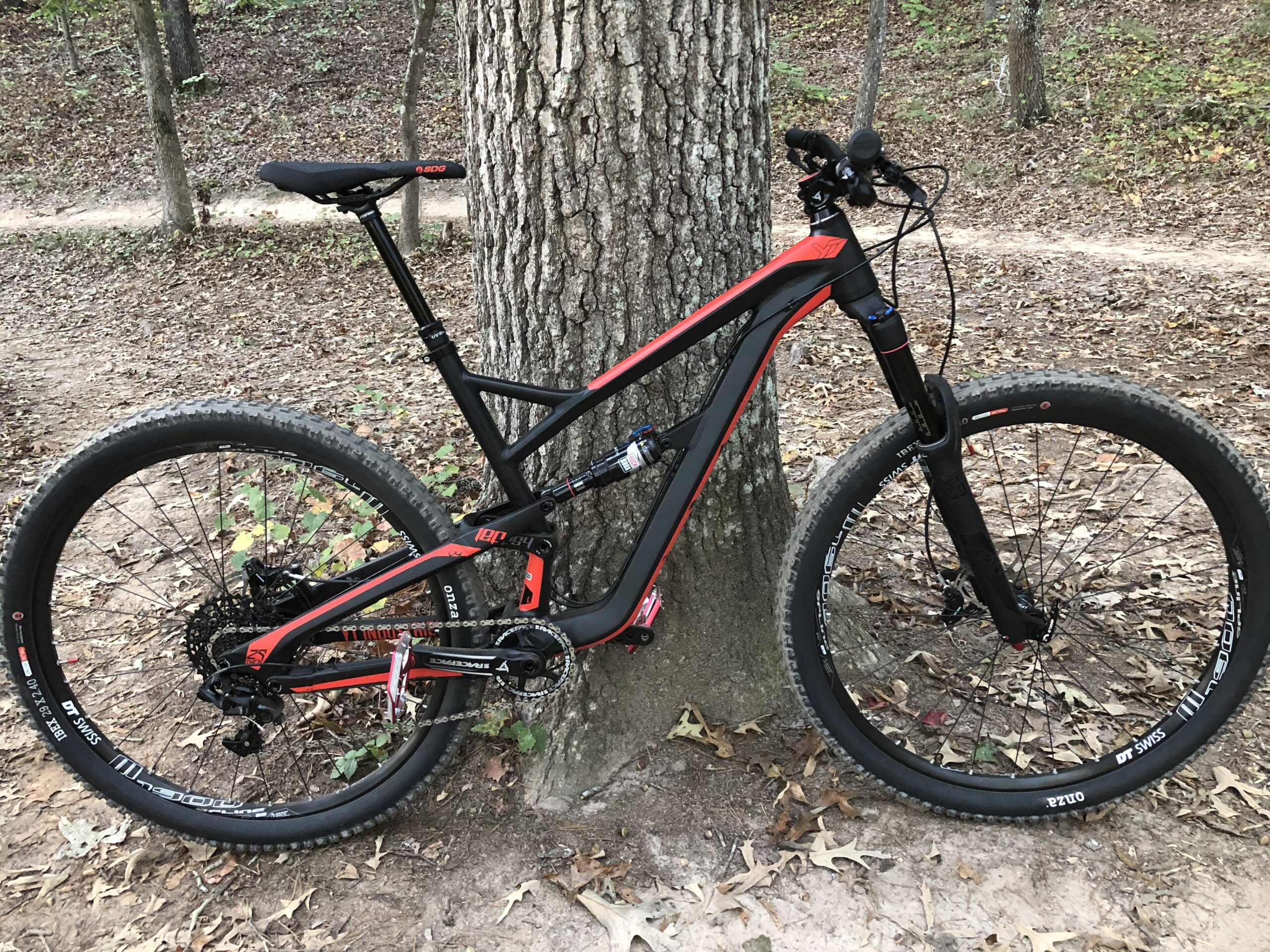 Yt industries Jeffsy: A mountain bike with a black and red frame is resting against a tree in a wooded area. The bike features wide tires and a modern design suitable for off-road trails, with a visible suspension fork and a dropper seat post. The background is covered in autumn leaves, hinting at a natural trail environment.