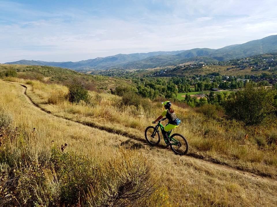 A person in bright athletic gear rides a mountain bike along a dirt trail surrounded by tall grass and shrubs. The landscape features rolling hills and distant mountains under a blue sky with wispy clouds, while a valley with trees and buildings is visible in the background. Emerald Mountain mountain bike trail.