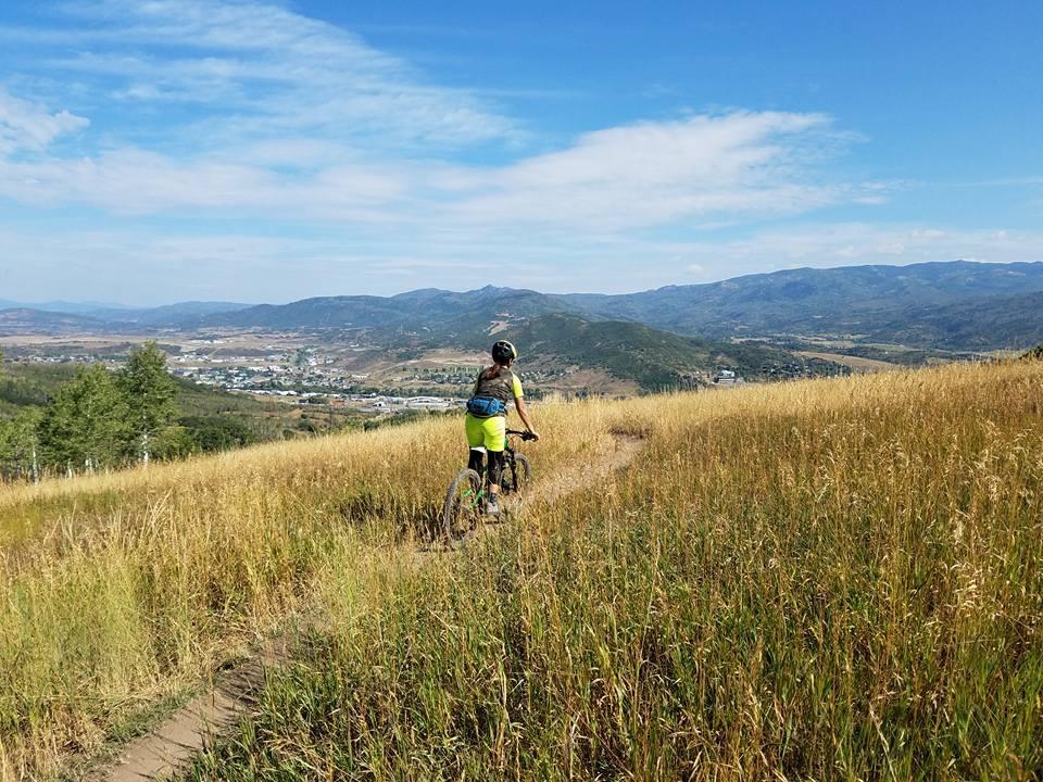 A person riding a mountain bike along a dirt trail through tall grass, with a scenic view of rolling hills and a small town in the distance underneath a blue sky with scattered clouds. Emerald Mountain mountain bike trail.