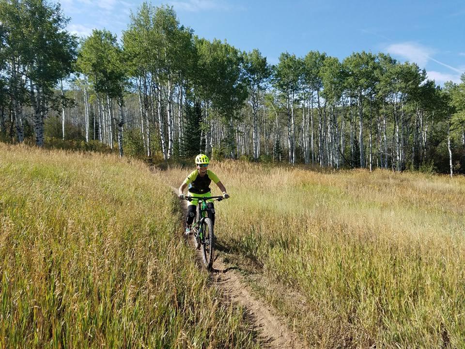 A person riding a mountain bike through a grassy field surrounded by trees. The cyclist is wearing a bright helmet and athletic gear, smiling as they navigate a dirt path. The background features a mix of green foliage and tall grasses under a clear blue sky. Emerald Mountain mountain bike trail.