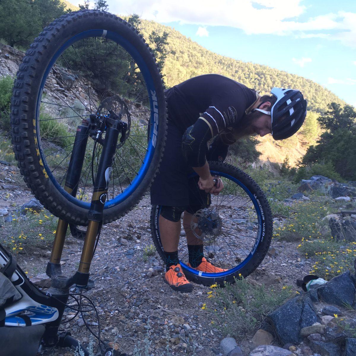 Michelin Wild AM: A mountain biker in a helmet is bending down to fix a flat tire on his bike in a scenic outdoor setting. In the foreground, a bike wheel is positioned upright, revealing its tire and components, while the rider is focused on the wheel on the ground. Surrounding them are rocky terrain and patches of wildflowers, with trees and hills in the background under a partly cloudy sky.
