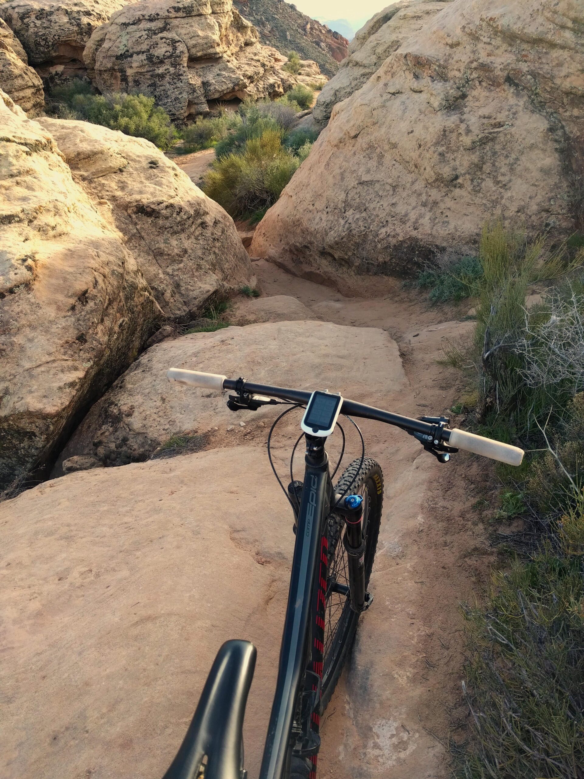 Specialized Epic Comp 29er: A mountain bike rests on a rocky trail, viewed from above the handlebars. The path is surrounded by large boulders and sparse vegetation, set against a backdrop of rolling hills and distant mountains. The sunlight casts a warm glow over the terrain.