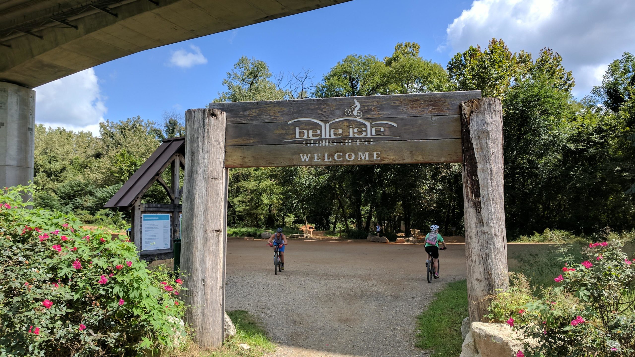 A wooden welcome sign for Belle Isle, situated under a bridge, with two cyclists entering the park. Lush greenery and flowering plants are visible around the entrance, creating a natural and inviting atmosphere. Belle Isle mountain bike trail.