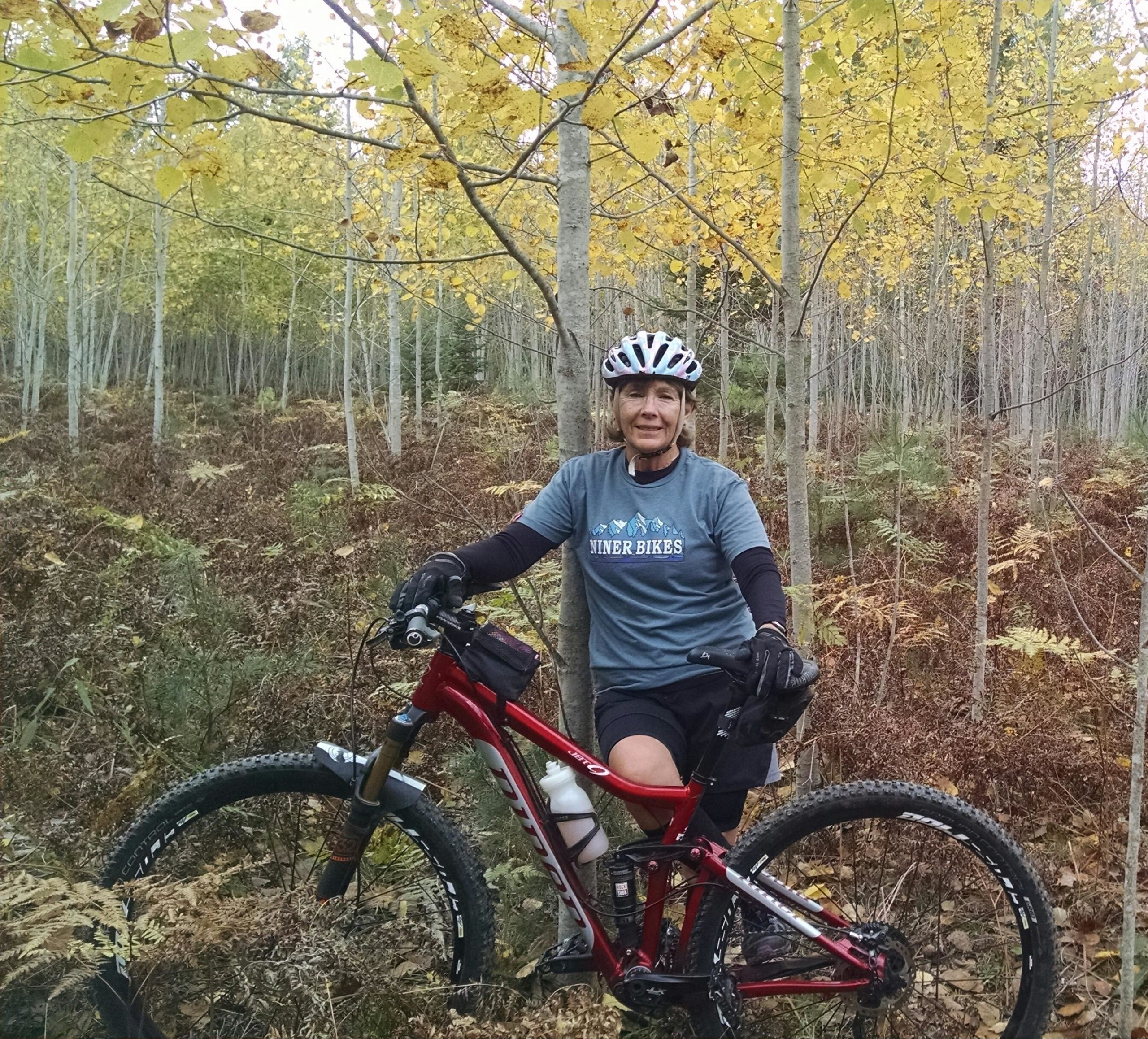 A smiling person stands next to a red mountain bike in a forested area during the fall, surrounded by yellow and orange foliage. They are wearing a gray T-shirt with "Niner Bikes" printed on it and a helmet. The background features tall trees and ferns, showcasing a natural and adventurous outdoor setting. Norway Ridge mountain bike trail.