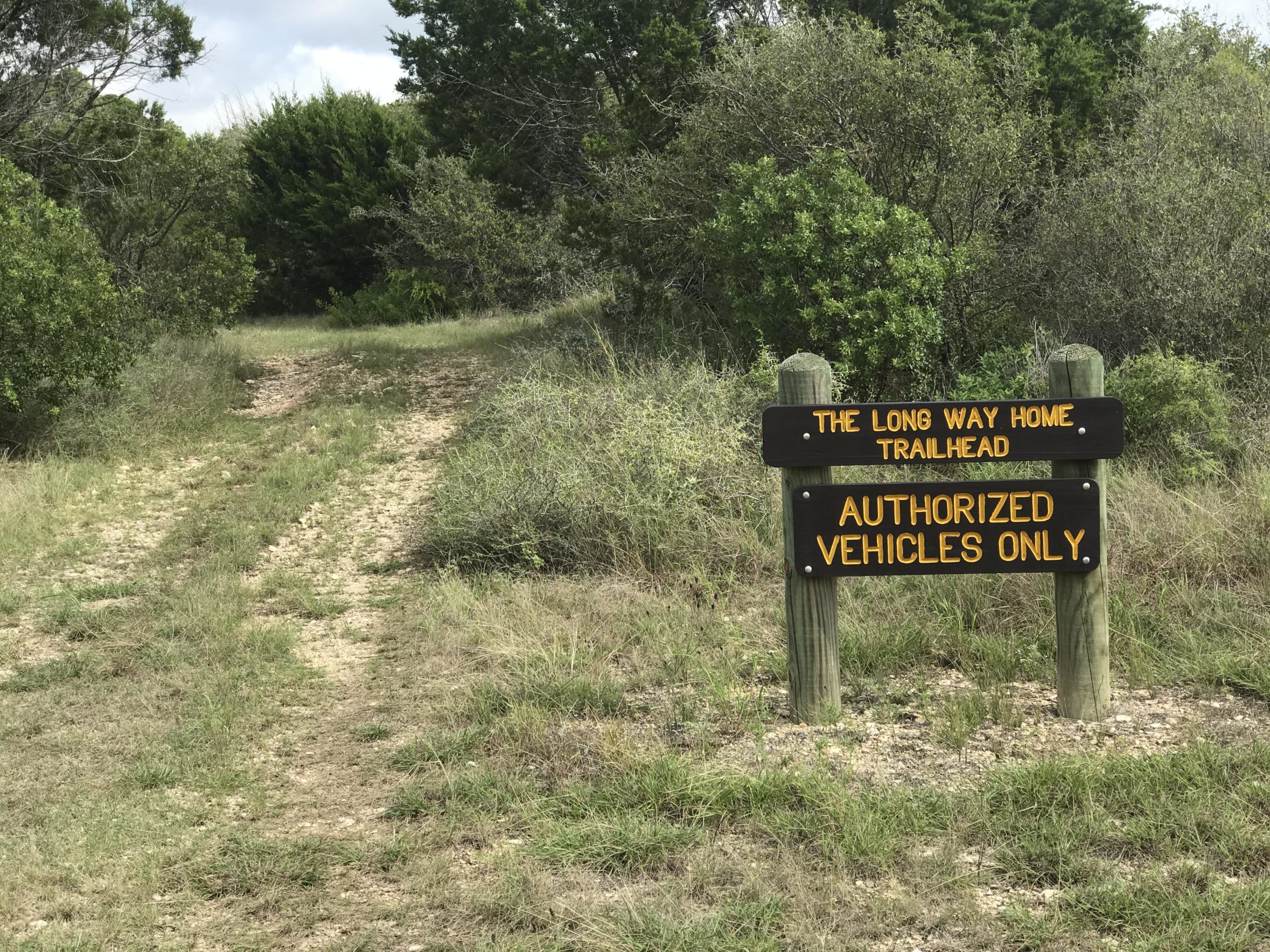 A dirt path leading into a wooded area, marked by a wooden sign that reads "The Long Way Home Trailhead" on the top and "Authorized Vehicles Only" on the bottom. Surrounding vegetation includes shrubs and grasses under a cloudy sky. Kickapoo Cavern State Park mountain bike trail.