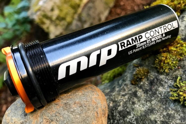 A close-up image of a black MRP Ramp Control suspension component resting on a rock, featuring an orange accent cap. The product label clearly displays the brand name "MRP," the model "RAMP CONTROL," and technical specifications, while a blurred natural background adds depth to the composition.