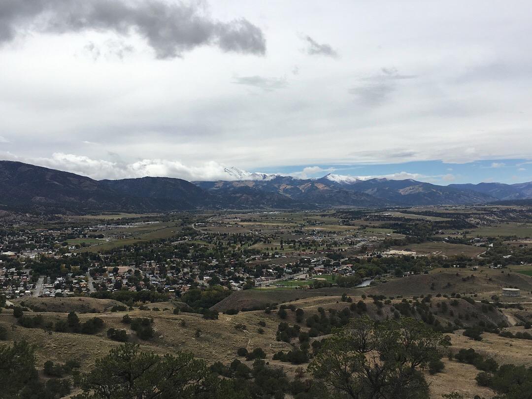 A scenic view of a valley surrounded by mountains, featuring a small town and lush fields. The sky is partly cloudy, with light clouds above the range of snow-capped peaks in the distance. Rusty Lung mountain bike trail.