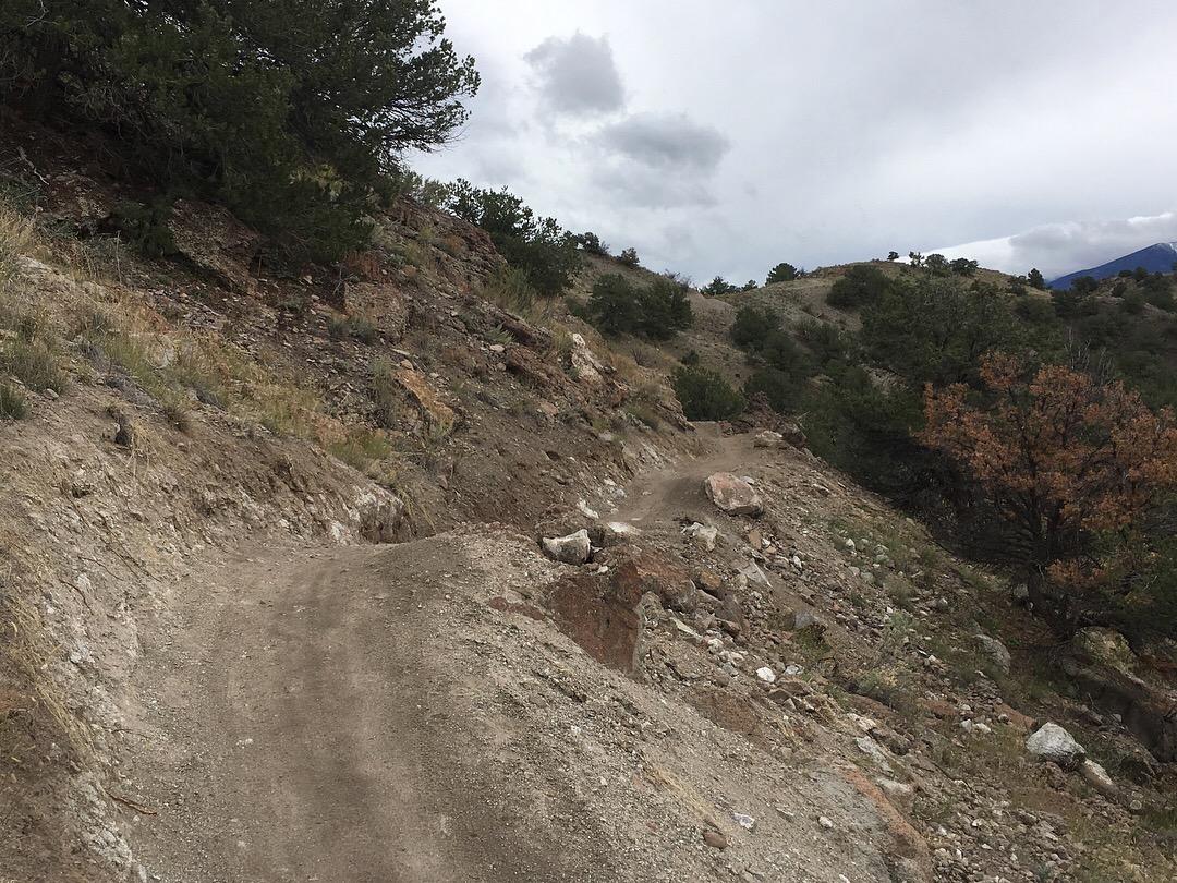 A narrow dirt trail winding along a hillside, surrounded by rocky terrain and sparse vegetation. The sky is overcast, with gray clouds looming overhead, and distant hills can be seen in the background. Rusty Lung mountain bike trail.
