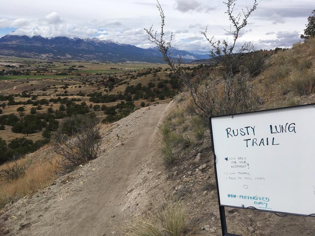 A dirt trail winding through a natural landscape, with a sign labeled "Rusty Lung Trail" indicating it is open for use. The sign advises to "know traffic" and "yield to small users," emphasizing that the trail is non-motorized. In the background, rolling hills and mountains can be seen under a cloudy sky. Rusty Lung mountain bike trail.