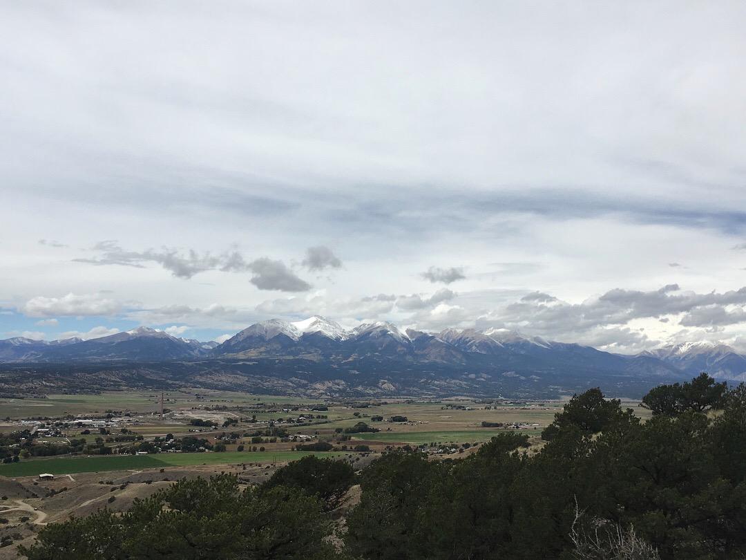 A scenic view of snow-capped mountains under a cloudy sky, with a valley below that features fields, small buildings, and patches of greenery. Rusty Lung mountain bike trail.