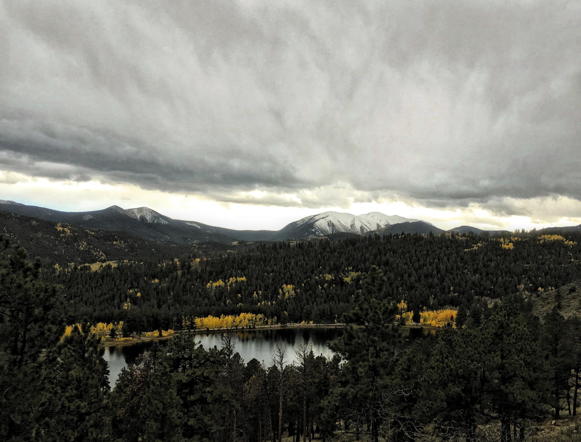 A mountainous landscape under a dramatic, overcast sky, featuring rolling hills covered in evergreen trees. A serene lake reflects the clouds, and patches of autumnal yellow foliage can be seen among the trees, adding color to the scene. Snow-capped mountains rise in the background, hinting at the elevation and beauty of the natural surroundings. Marshall Pass Road / #200 / #203 / #243 mountain bike trail.