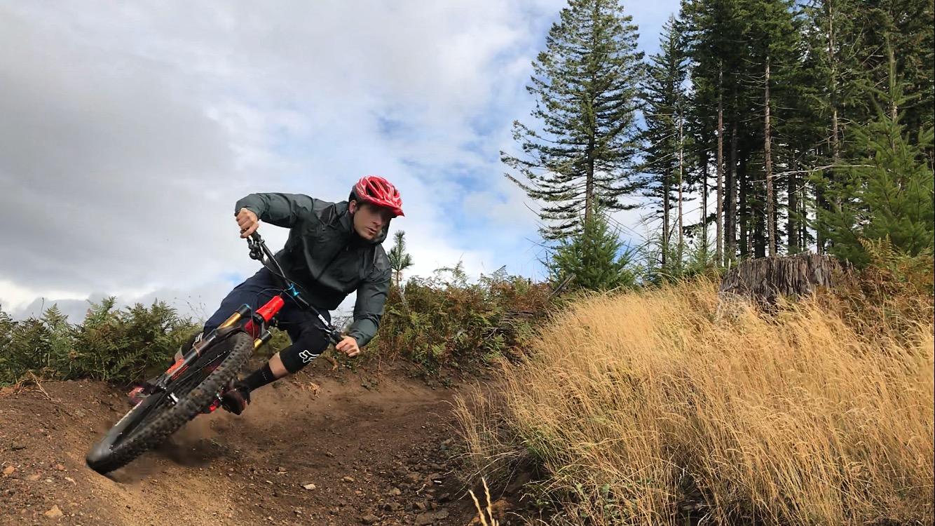 A mountain biker leans into a turn on a dirt trail surrounded by tall grass and trees, set against a cloudy sky. The cyclist wears a red helmet and a dark jacket, showcasing an action-packed moment in outdoor sports. Cold Creek mountain bike trail.