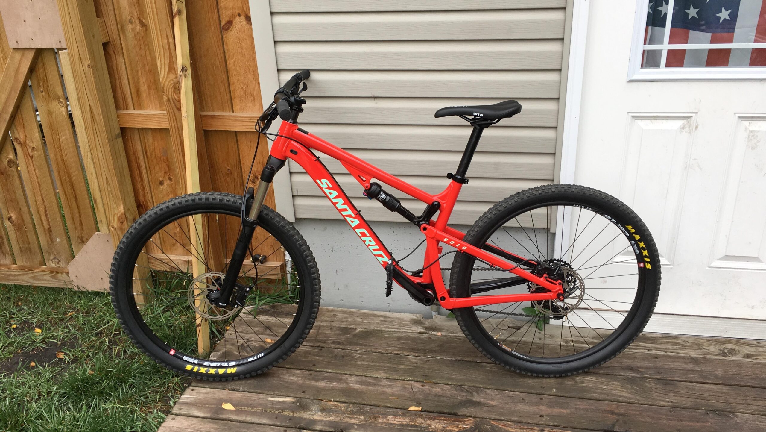 Santa Cruz 5010: A vibrant red mountain bike from Santa Cruz, parked on a wooden deck. The bike features a sturdy frame, rugged tires, and front suspension. In the background, there is wooden fencing and a door with a partially visible American flag.