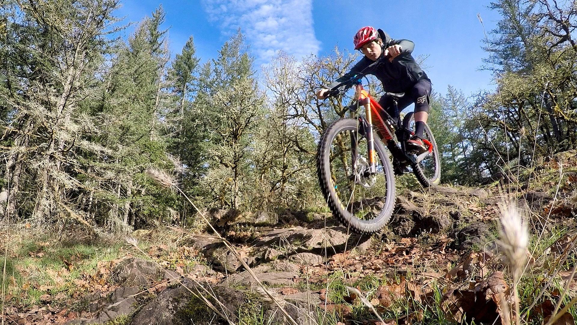 A mountain biker in a red helmet and black attire jumps over rocky terrain in a forested area, with green trees and blue skies in the background. Round Lake mountain bike trail.