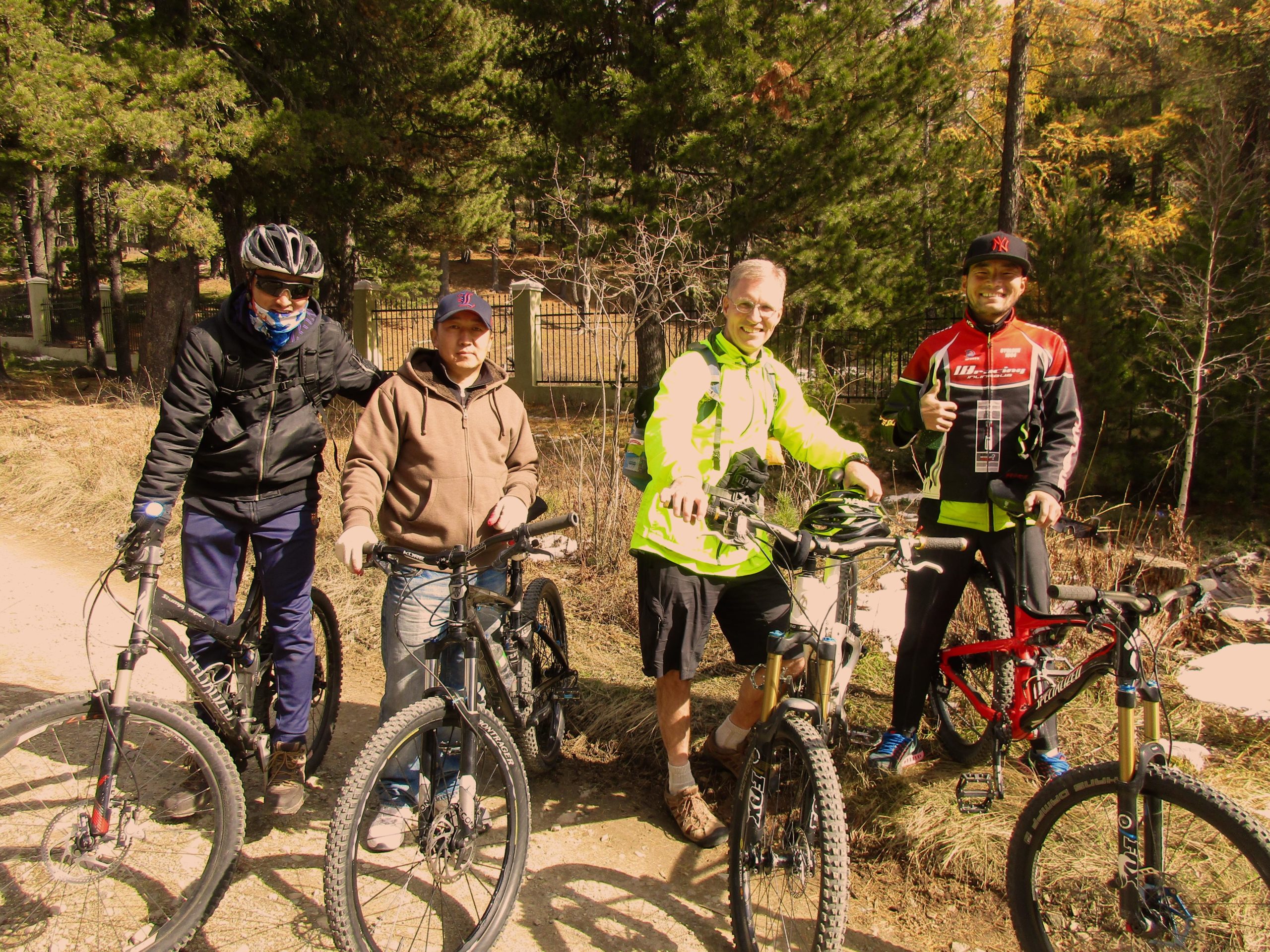 Four mountain bikers pose together on a dirt path surrounded by trees. They are wearing a mix of cycling gear, including helmets and jackets. The group includes a man in a bright green jacket, another with a red and black jersey, and two others in casual attire, all standing next to their mountain bikes and smiling. The background features greenery and foliage, indicating an outdoor setting. Shargamorit Khandgait Hills East mountain bike trail.