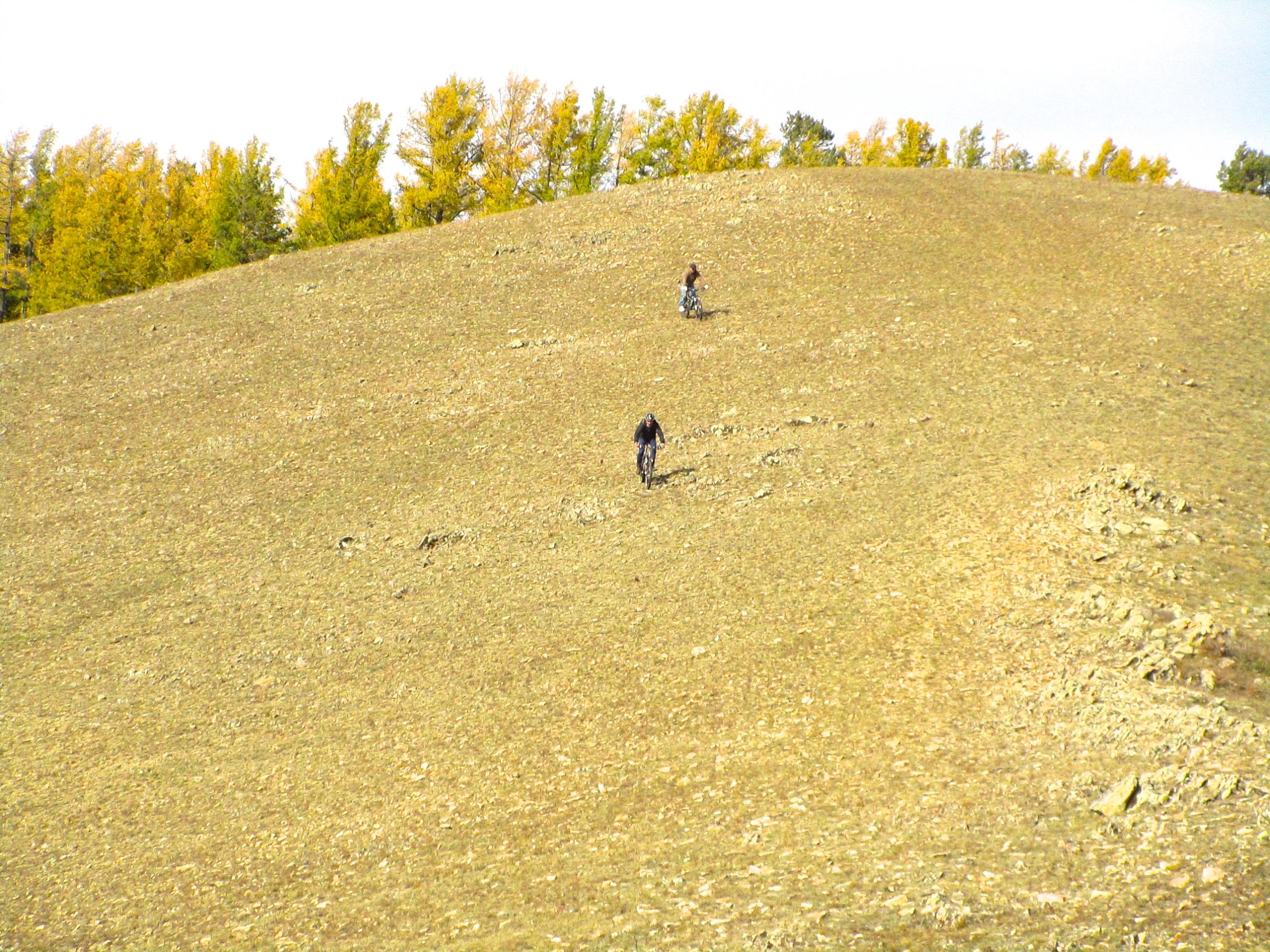 Two mountain bikers riding up a sloped terrain with scattered rocks, surrounded by trees displaying autumn colors in the background. The sky is bright and clear. Selkh Am mountain bike trail.