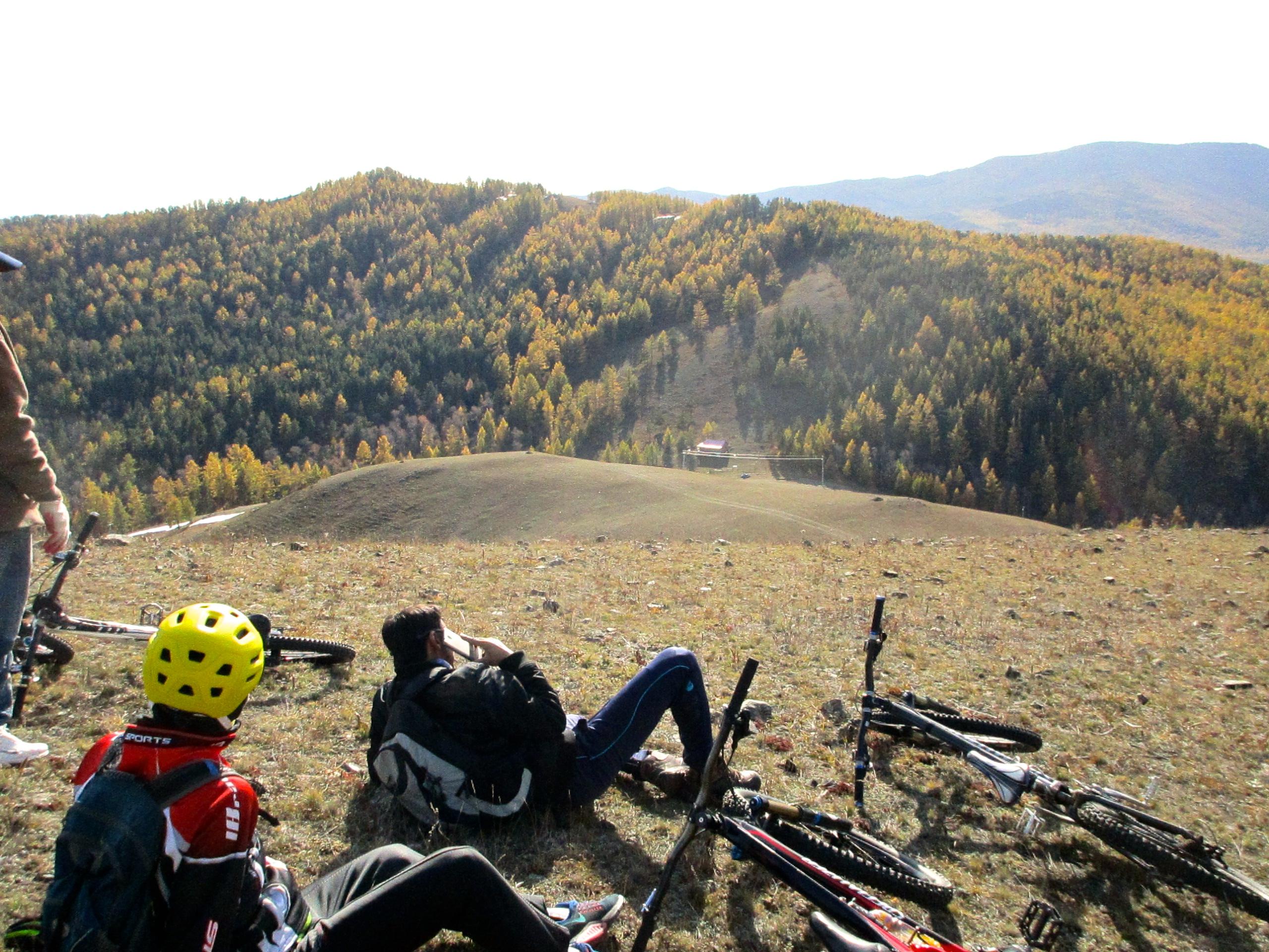 A scenic view of a mountainous landscape with colorful autumn foliage. In the foreground, two mountain bikers relax on a grassy slope, one sitting and using a phone, while the other lies back. Several bicycles are positioned nearby. The background features rolling hills and trees with golden leaves, under a clear sky. Selkh Am mountain bike trail.