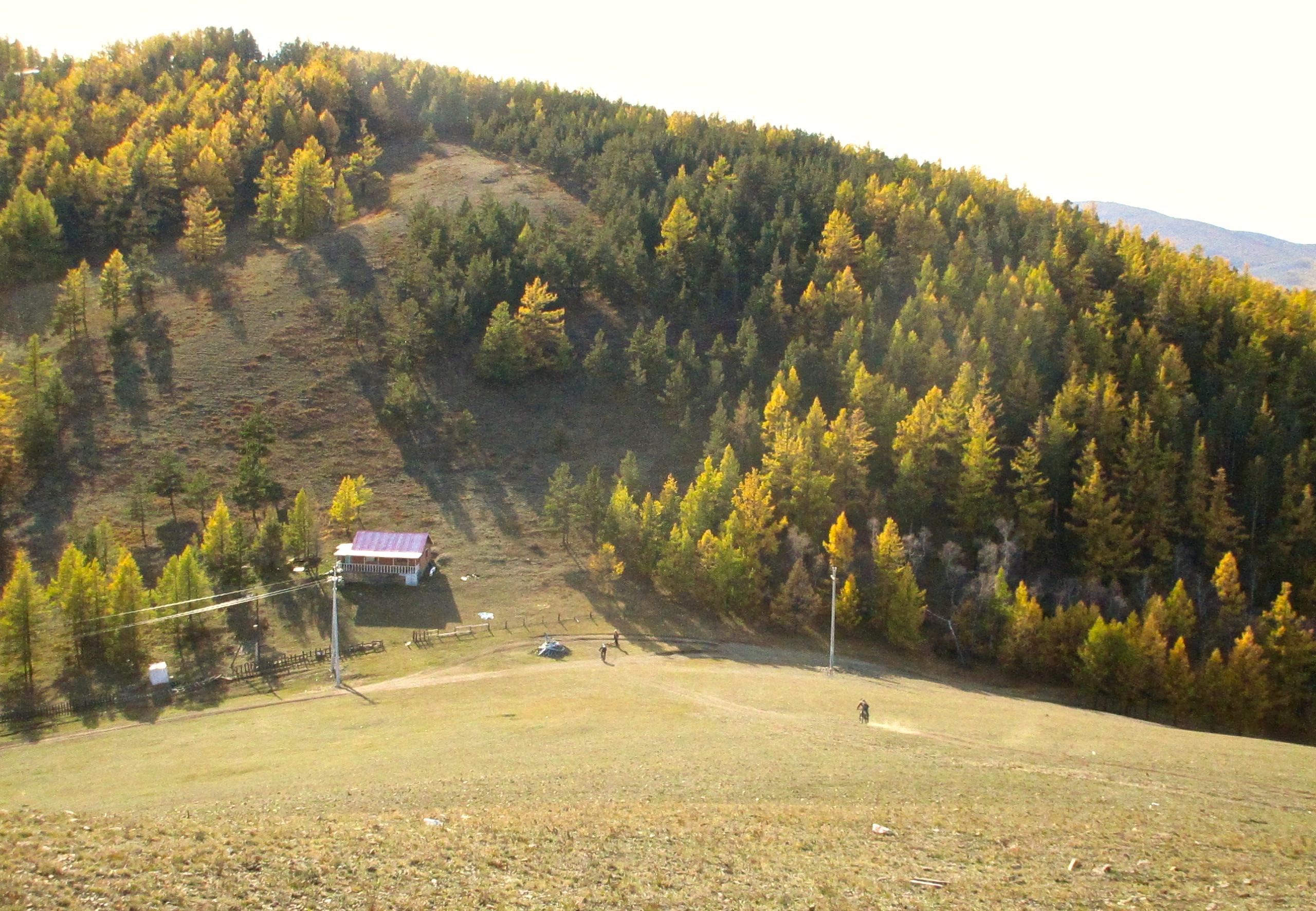 A picturesque landscape featuring a small, rustic house with a red roof nestled in a valley surrounded by rolling hills. The hills are adorned with lush green and golden trees, indicating the onset of autumn. Sunlight casts long shadows across the terrain, enhancing the tranquility of the scene. In the foreground, a lone figure can be seen walking along a path, adding a sense of scale to the expansive natural setting. Selkh Am mountain bike trail.