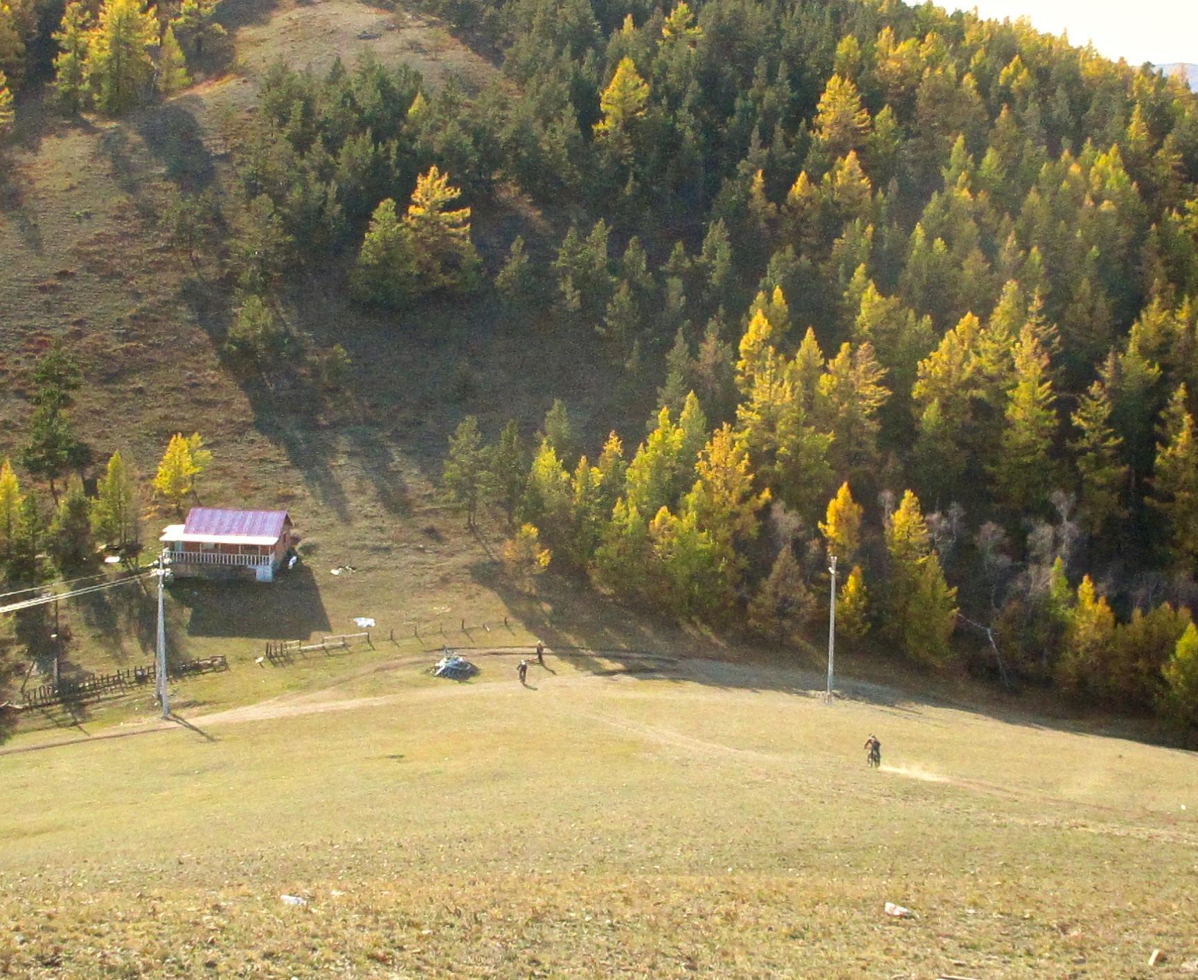 A scenic view of a hilly landscape featuring a small cabin with a red roof, surrounded by trees displaying autumn colors. A person rides a bicycle on a dirt path in the foreground, while a power line and fence are visible near the cabin. The hills are partly shaded, creating a contrast between light and shadow. Selkh Am mountain bike trail.