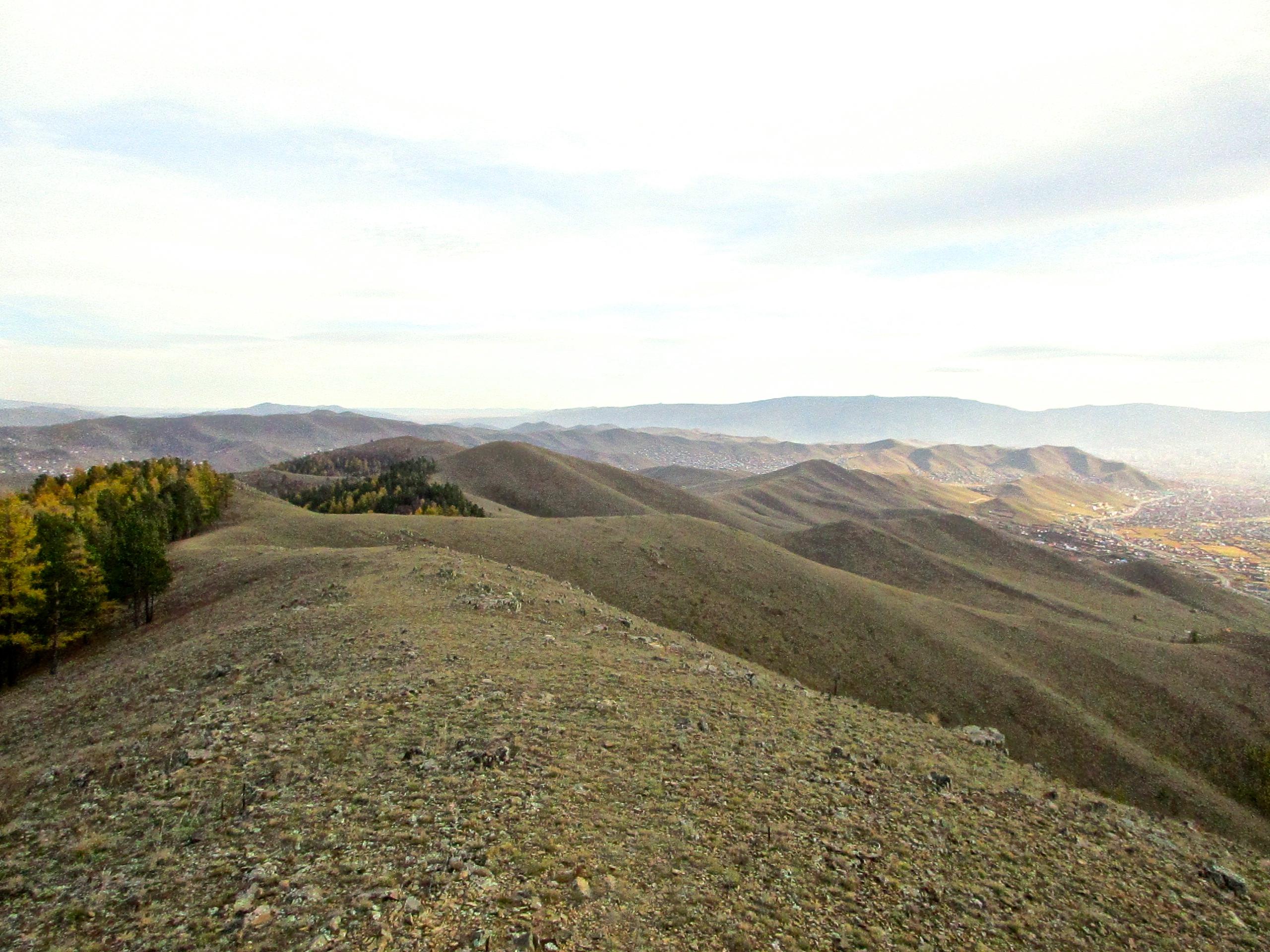 A panoramic view of rolling hills and mountains under a cloudy sky, with a patch of trees in the foreground displaying autumn colors. The landscape features rocky terrains and valleys stretching into the distance, evoking a serene and natural environment. Selkh Am mountain bike trail.