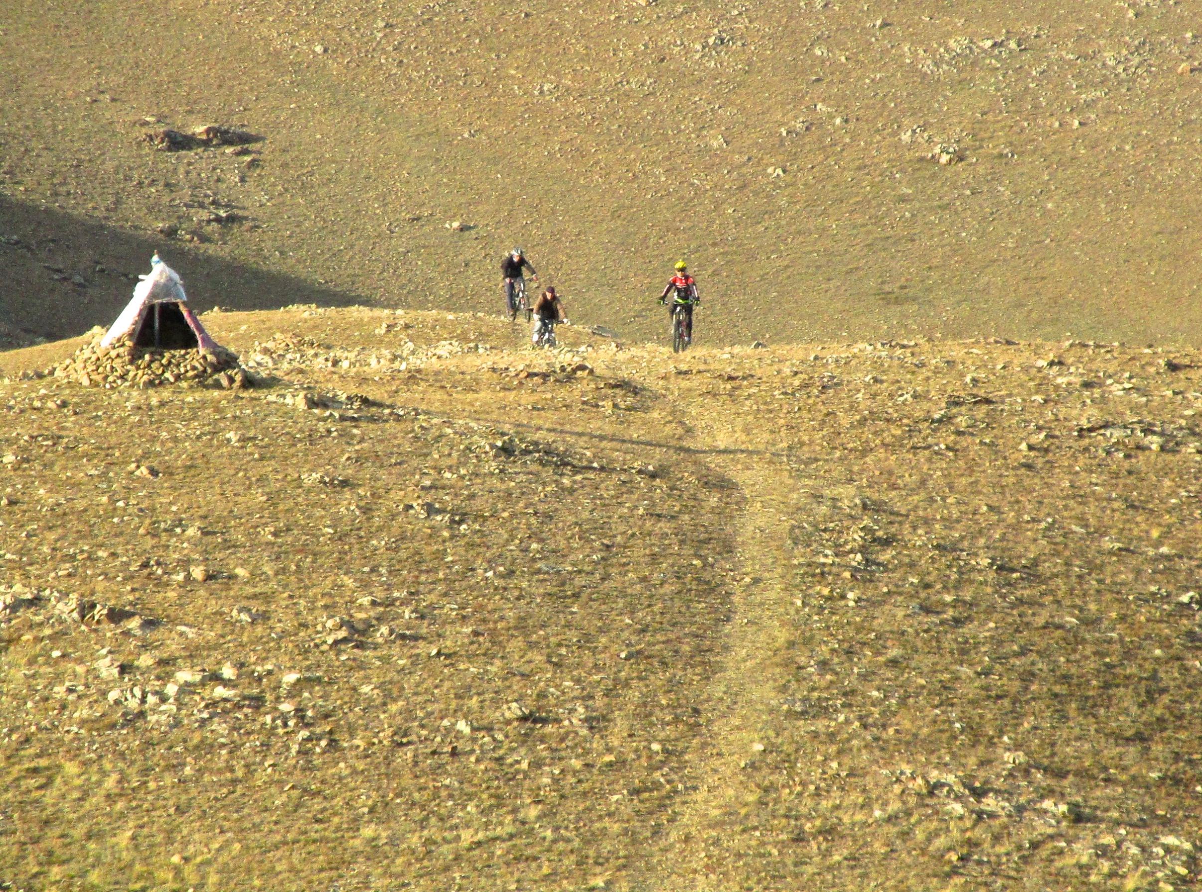 Three mountain bikers riding on a rocky path in a barren landscape, with a small tent structure made of rocks and fabric visible on the left. The terrain is dry and hilly, under clear skies. Selkh Am mountain bike trail.
