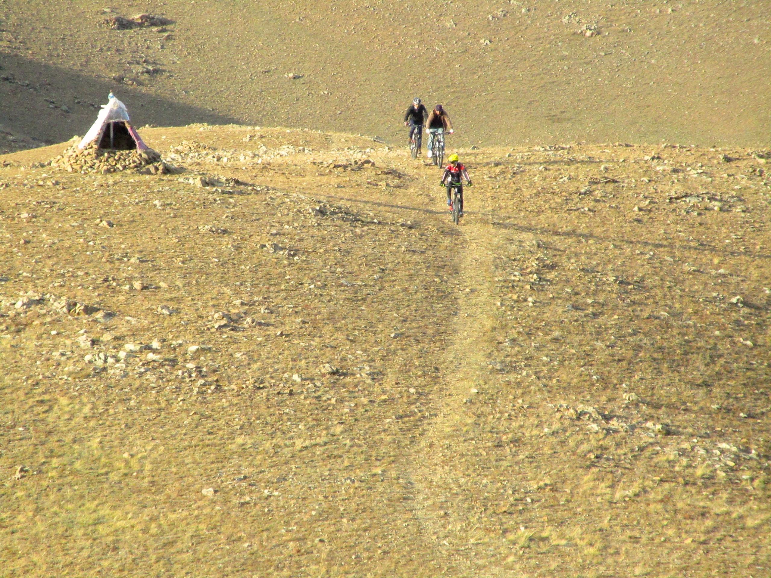 A group of three mountain bikers riding along a narrow dirt path across a barren landscape, with a small tent made of stones and fabric visible in the background. The terrain is rocky and devoid of vegetation, under a clear sky. Selkh Am mountain bike trail.