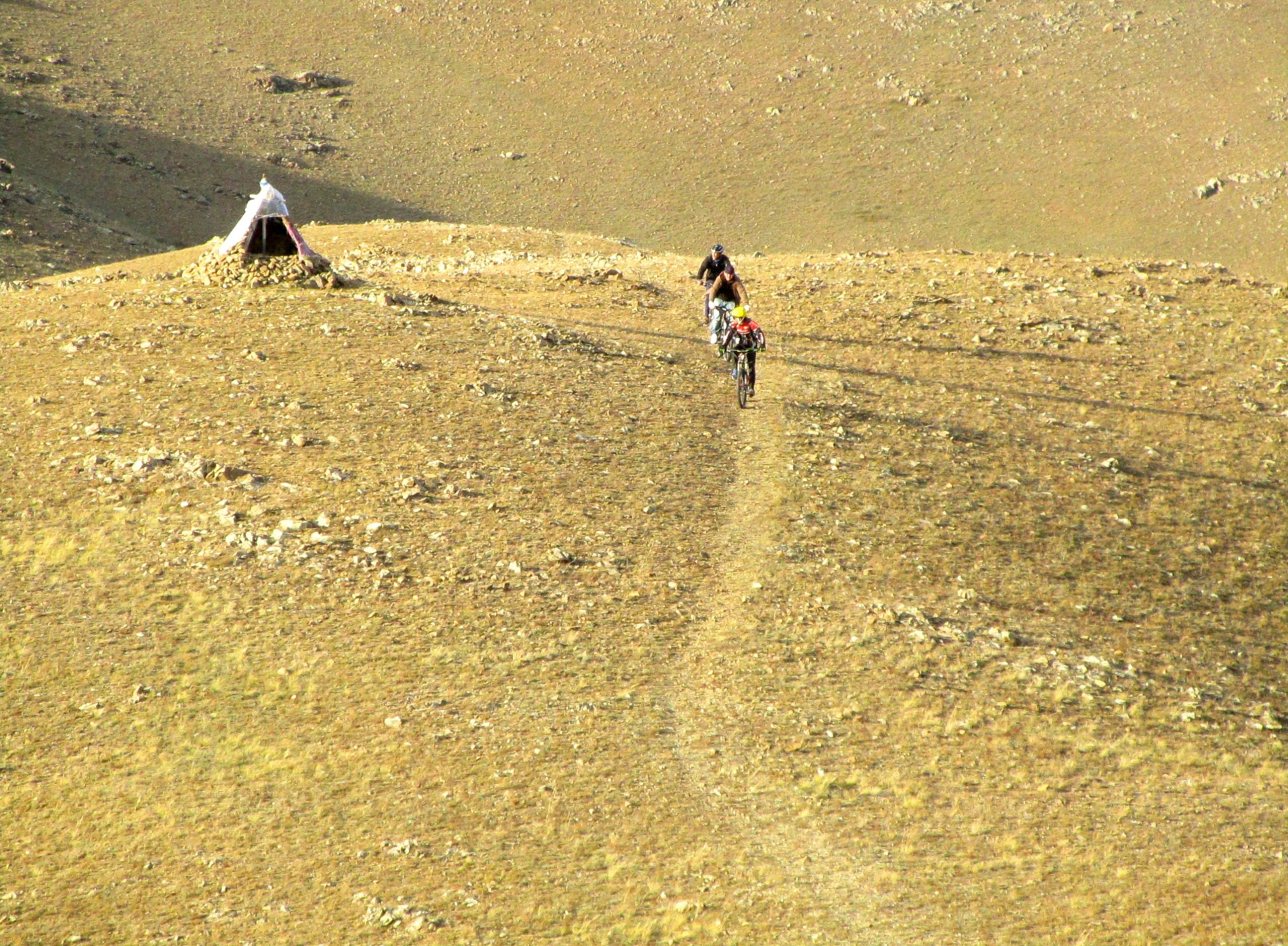 Two cyclists riding on a rocky trail in a mountainous landscape, with a small tent made of stones visible in the background. The area appears barren and dry, under a clear sky. Selkh Am mountain bike trail.
