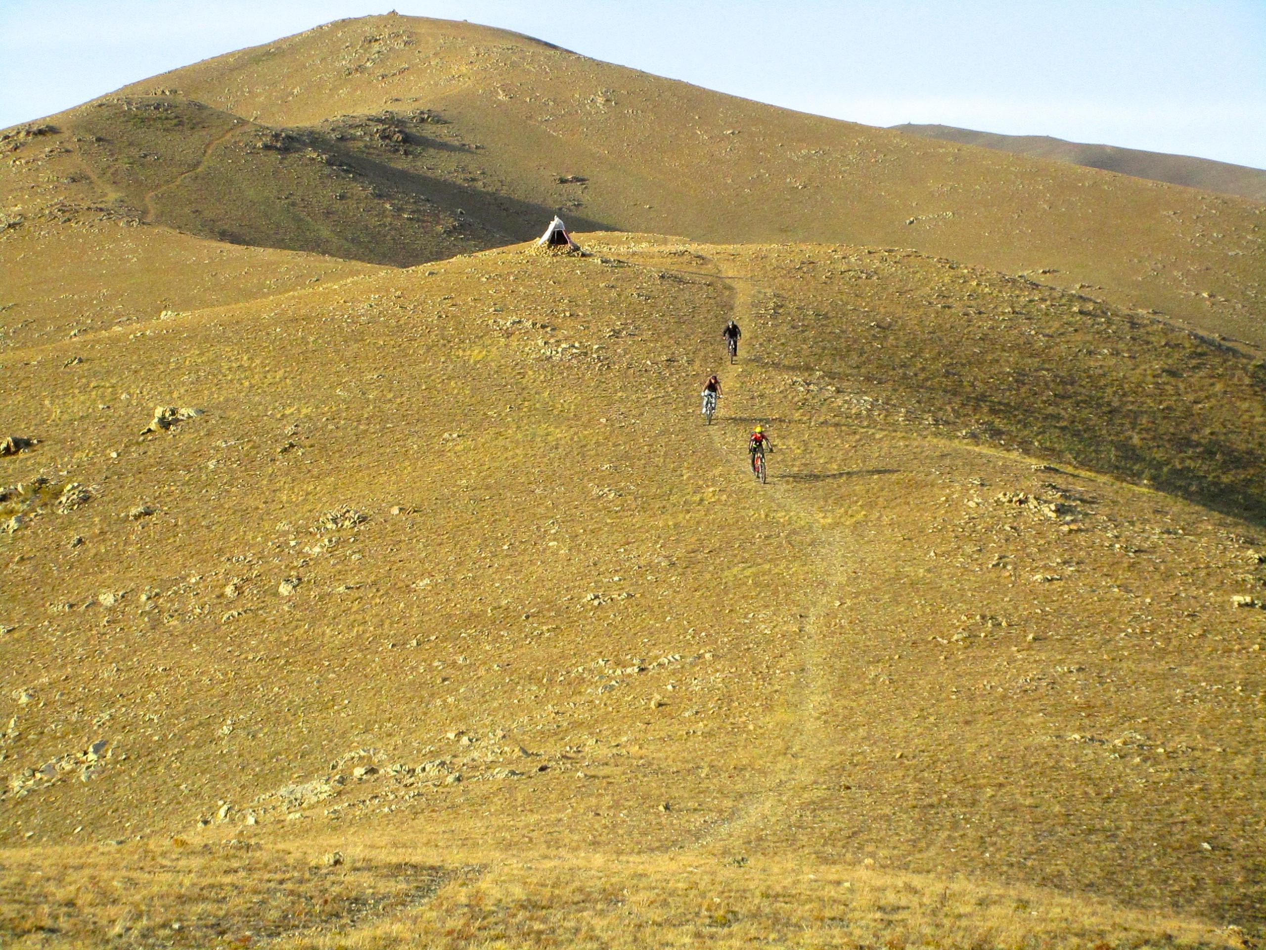 Three mountain bikers navigate a winding trail on a hillside covered with golden grass and scattered rocks, with a small tent visible in the distance against a backdrop of rolling hills. Selkh Am mountain bike trail.