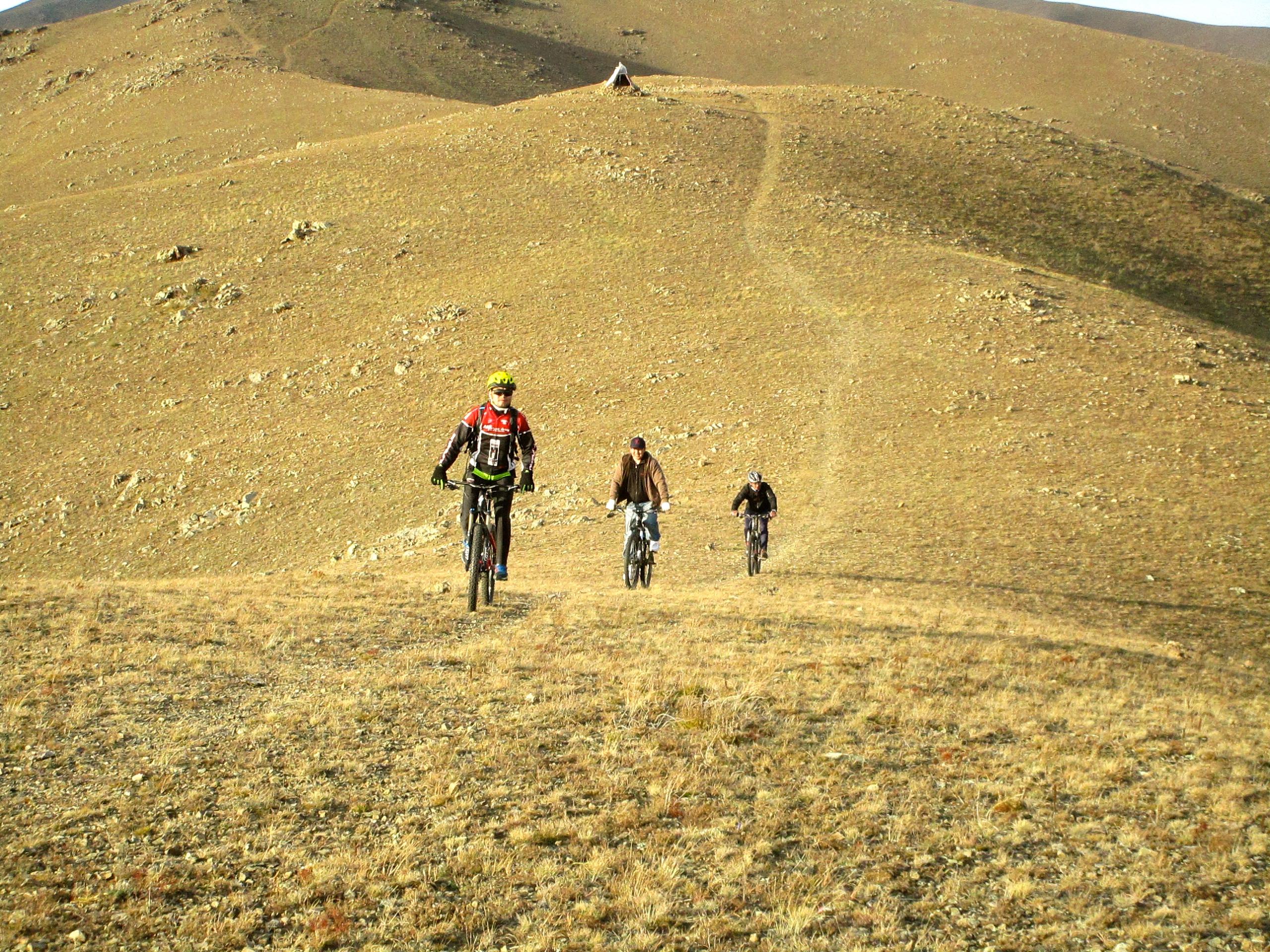 Three mountain bikers climbing a grassy slope, with rolling hills in the background. The landscape is primarily dry and rocky, featuring minimal vegetation. A small structure can be seen at the top of the hill. Selkh Am mountain bike trail.