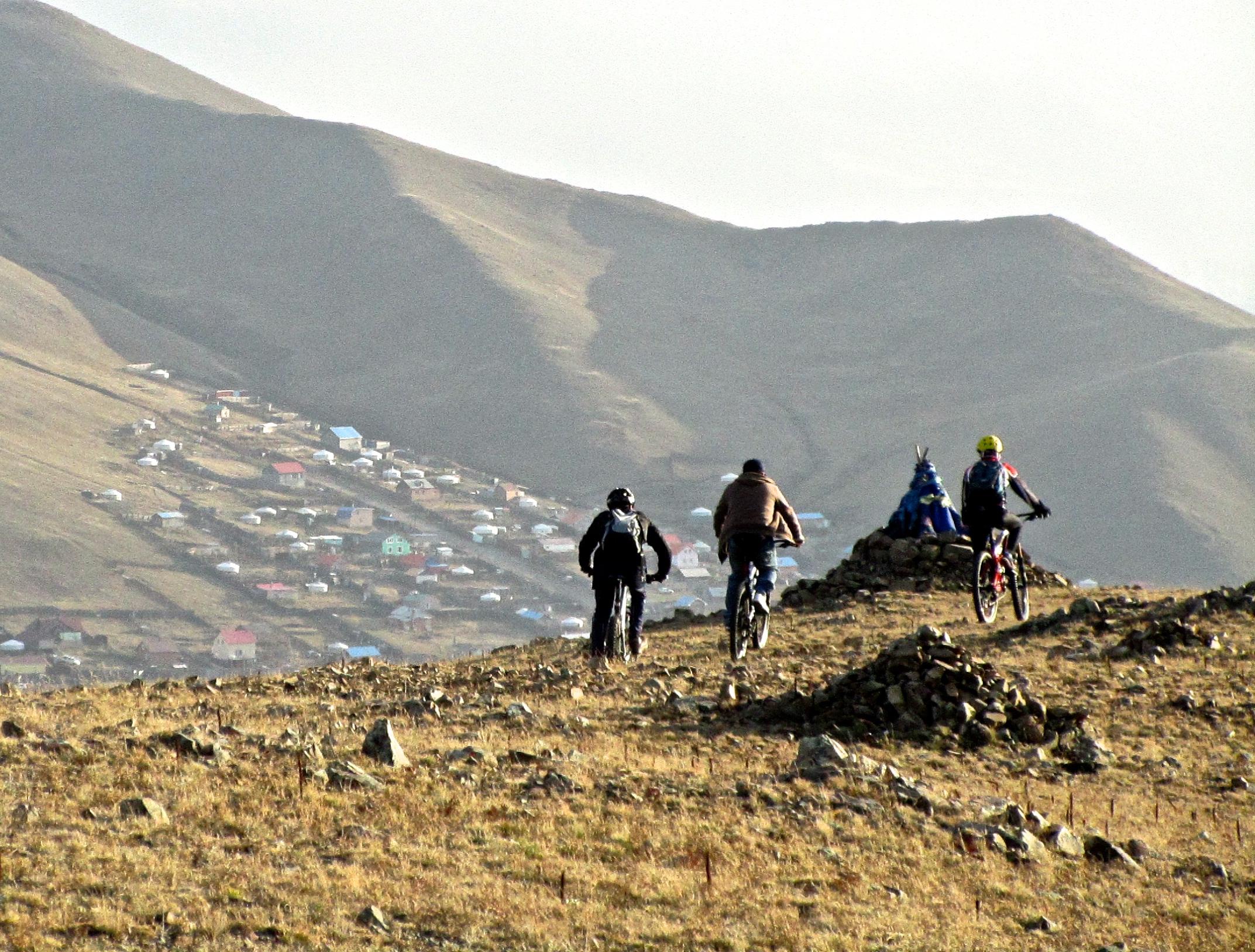 Four mountain bikers ride along a grassy hillside, with distant mountains and a small village visible below. The bikers are positioned away from the camera, showcasing their movement across the uneven terrain. In the foreground, a stone pile is visible, adding a natural element to the rugged landscape. The sky is slightly overcast, creating a serene and adventurous atmosphere. Selkh Am mountain bike trail.