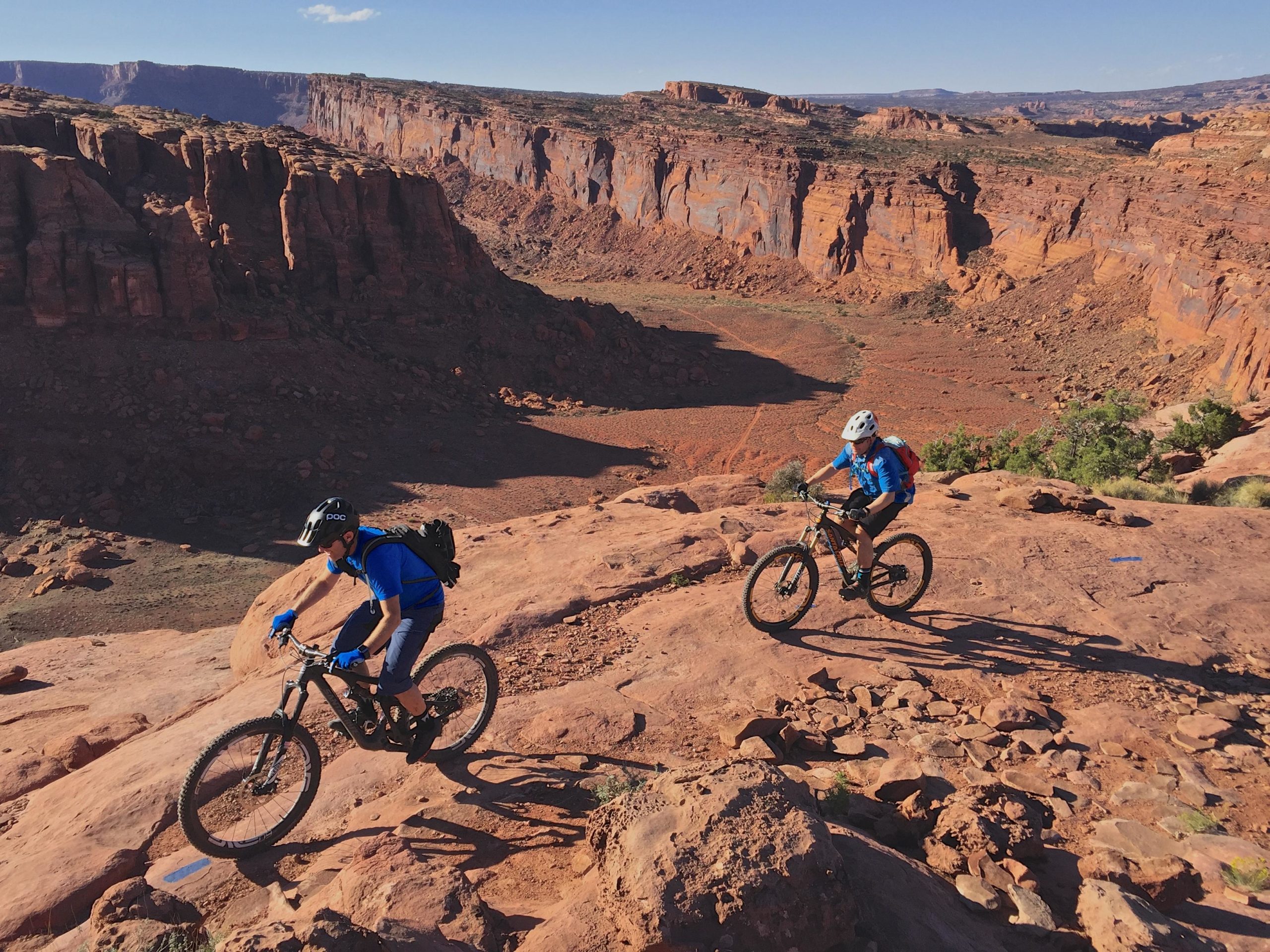 Two mountain bikers in blue attire navigate rough, rocky terrain in a canyon landscape. The red rock formations rise steeply in the background, under a clear blue sky. Captain Ahab mountain bike trail.