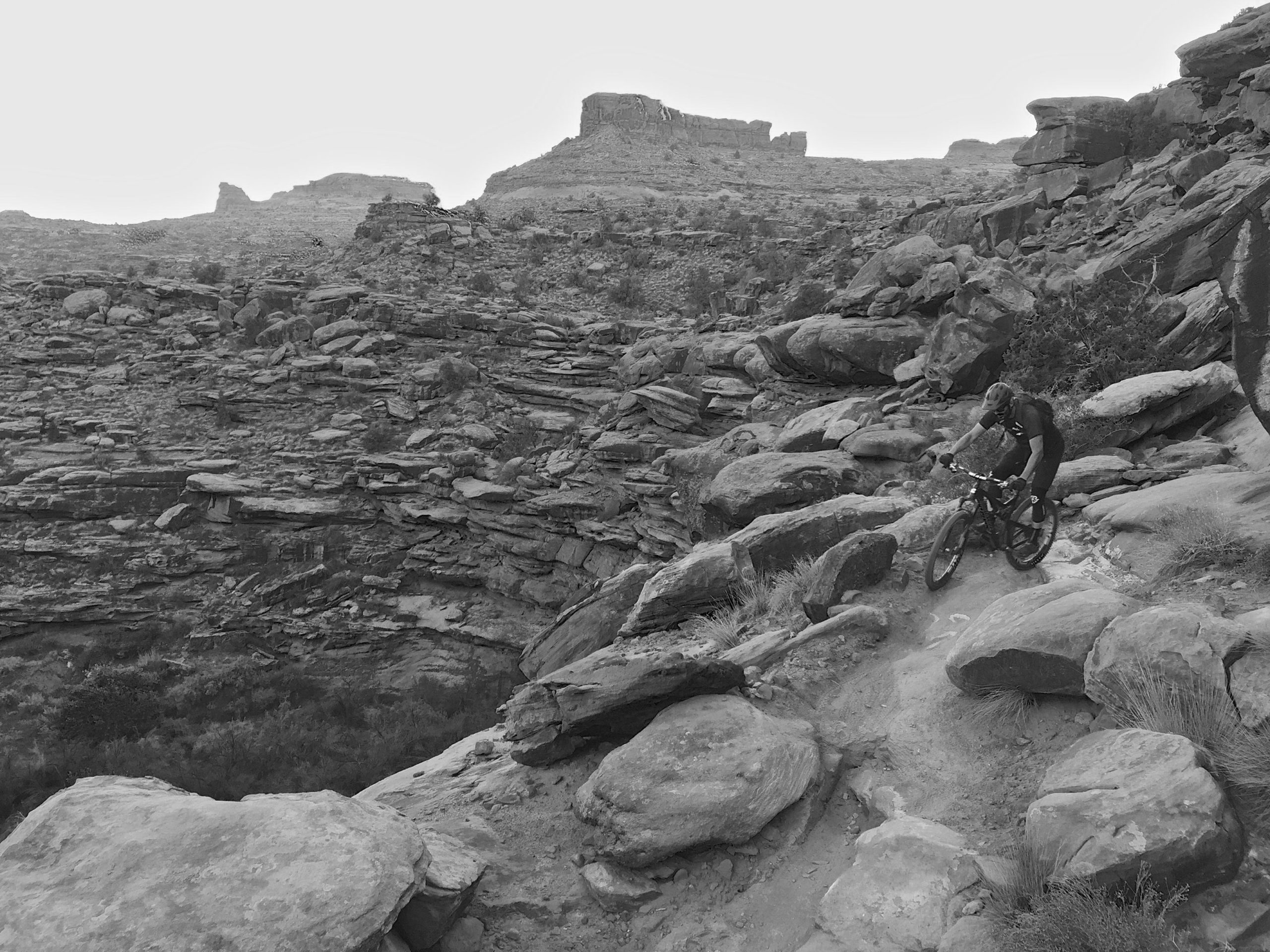 A mountain biker navigating a rocky trail surrounded by rugged terrain, captured in black and white. The dramatic landscape features large boulders and steep cliffs in the background, highlighting the challenging environment for outdoor adventure. Captain Ahab mountain bike trail.
