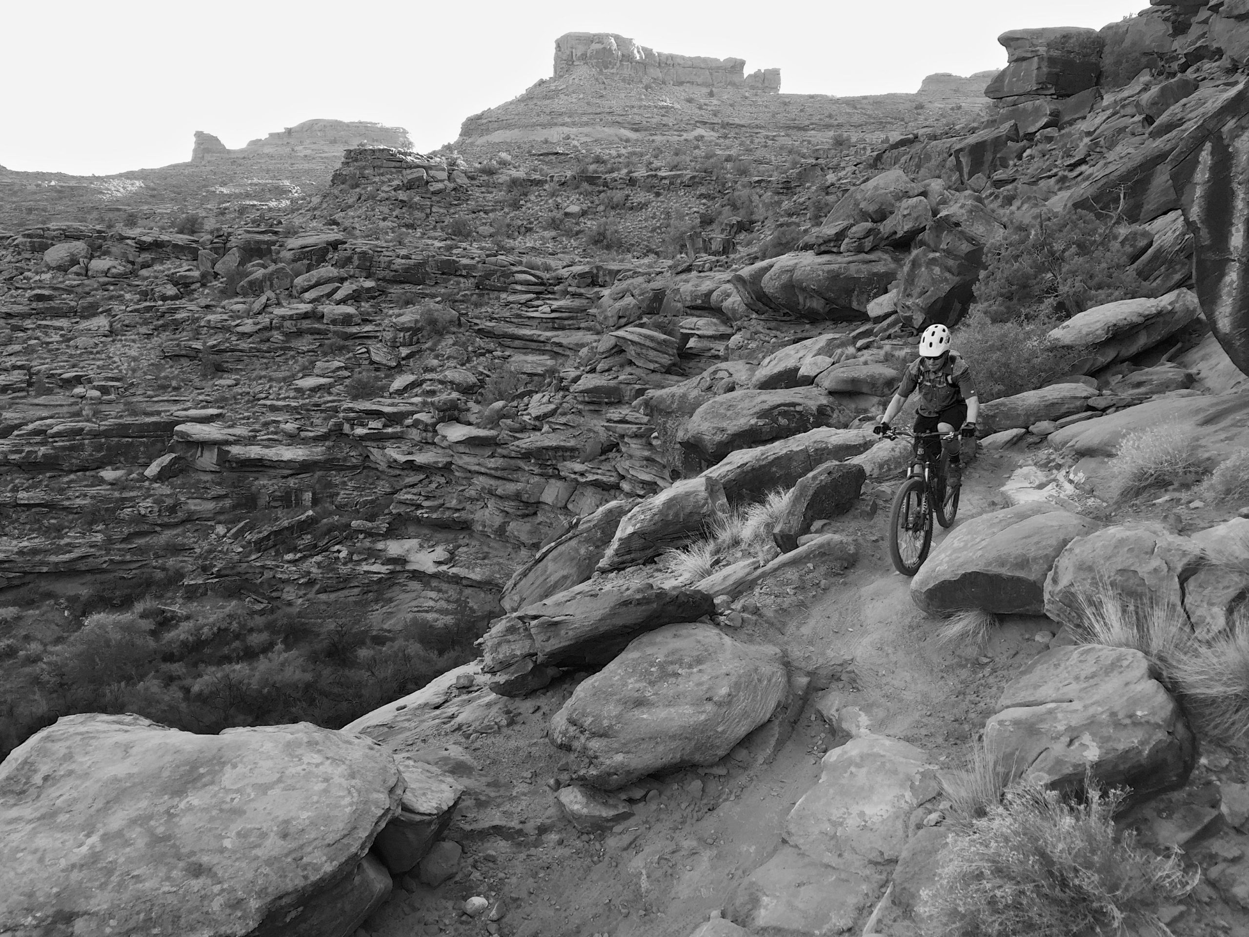 A child riding a mountain bike along a rocky trail with steep canyon walls in the background, surrounded by rugged terrain and sparse vegetation. The image is in black and white. Captain Ahab mountain bike trail.