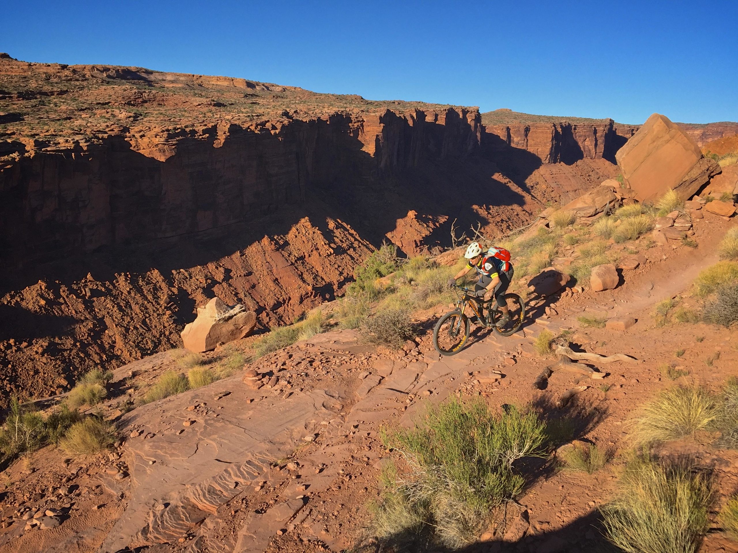 A mountain biker navigating a rocky trail on a canyon ridge under a clear blue sky, surrounded by steep red rock formations and sparse vegetation. Porcupine Rim mountain bike trail.