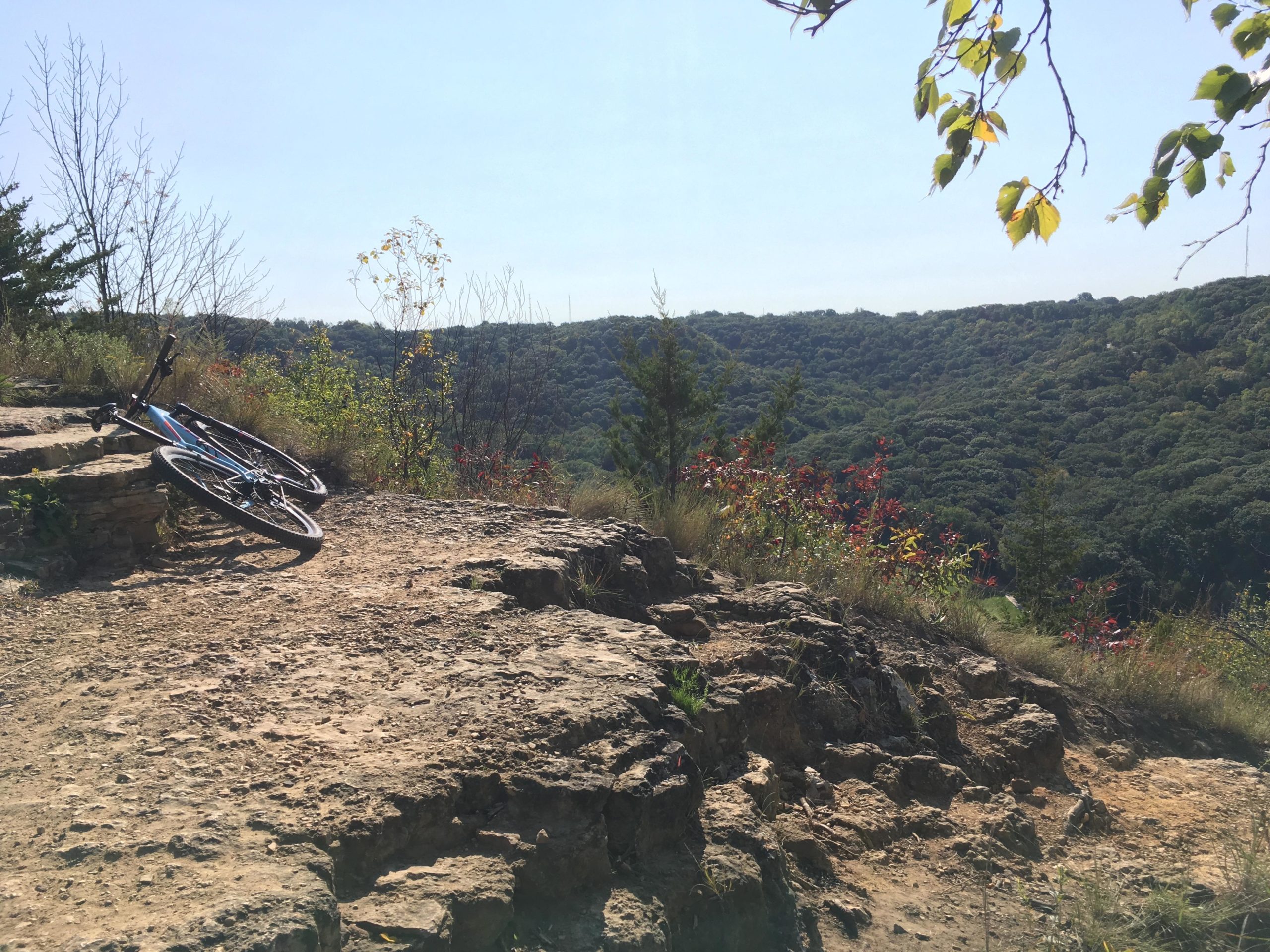 A mountain bike resting on rocky terrain with a scenic view of a lush, green valley and trees in the distance under a clear blue sky. Rotary Vista mountain bike trail.