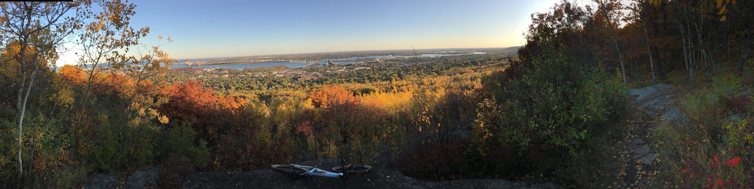 A panoramic view of a vibrant autumn landscape from a mountain overlook, showcasing a mix of colorful foliage in shades of orange, red, and yellow. In the foreground, a mountain bike is resting on a rock, while the background features a serene river winding through the valley, with a town visible among the trees under a clear blue sky. Piedmont mountain bike trail.