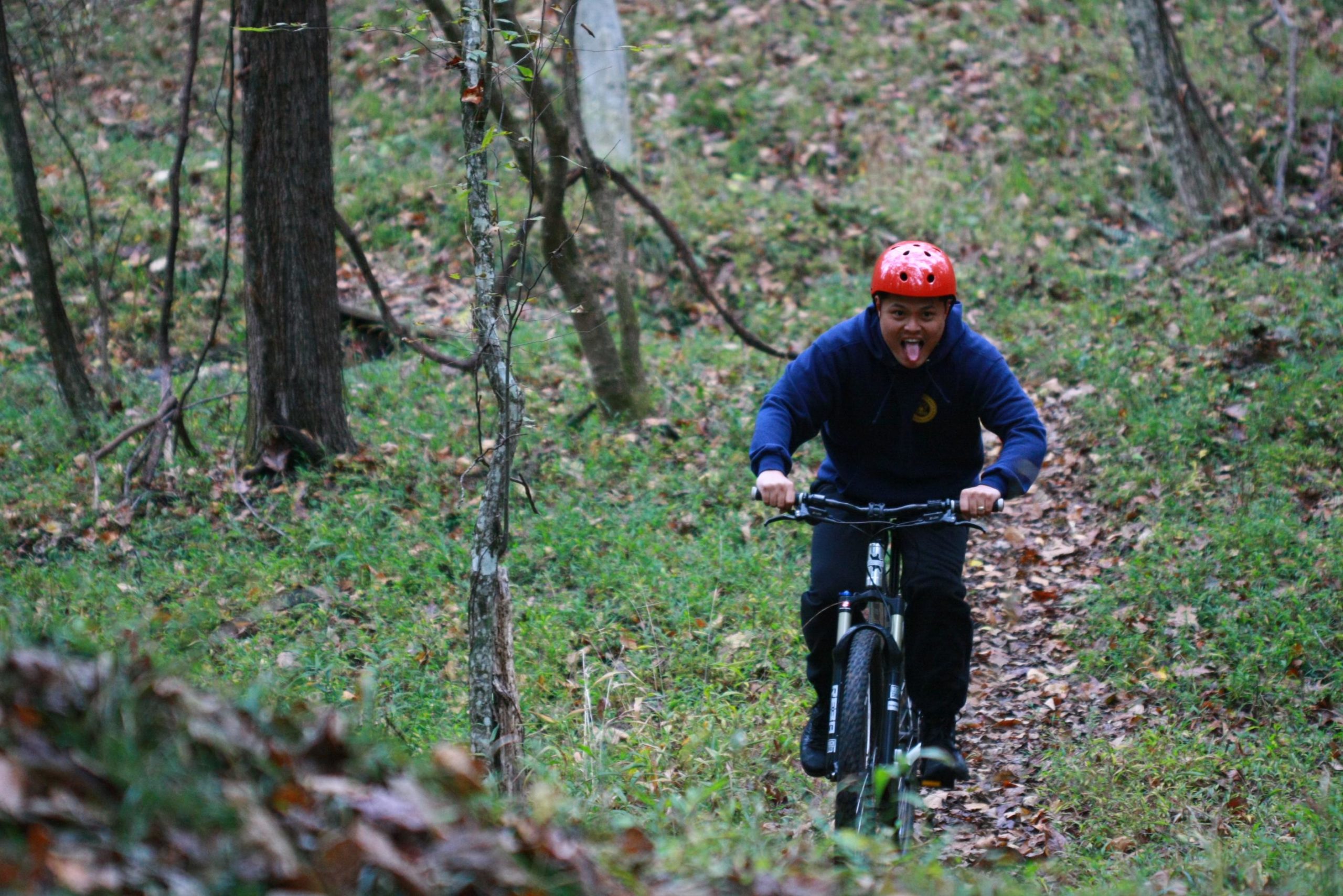 A person wearing a red helmet and a navy hoodie is riding a mountain bike on a wooded trail, sticking out their tongue playfully. The background features lush green grass and scattered autumn leaves, with trees lining the path. Cedar Ridge Chatmoss mountain bike trail.