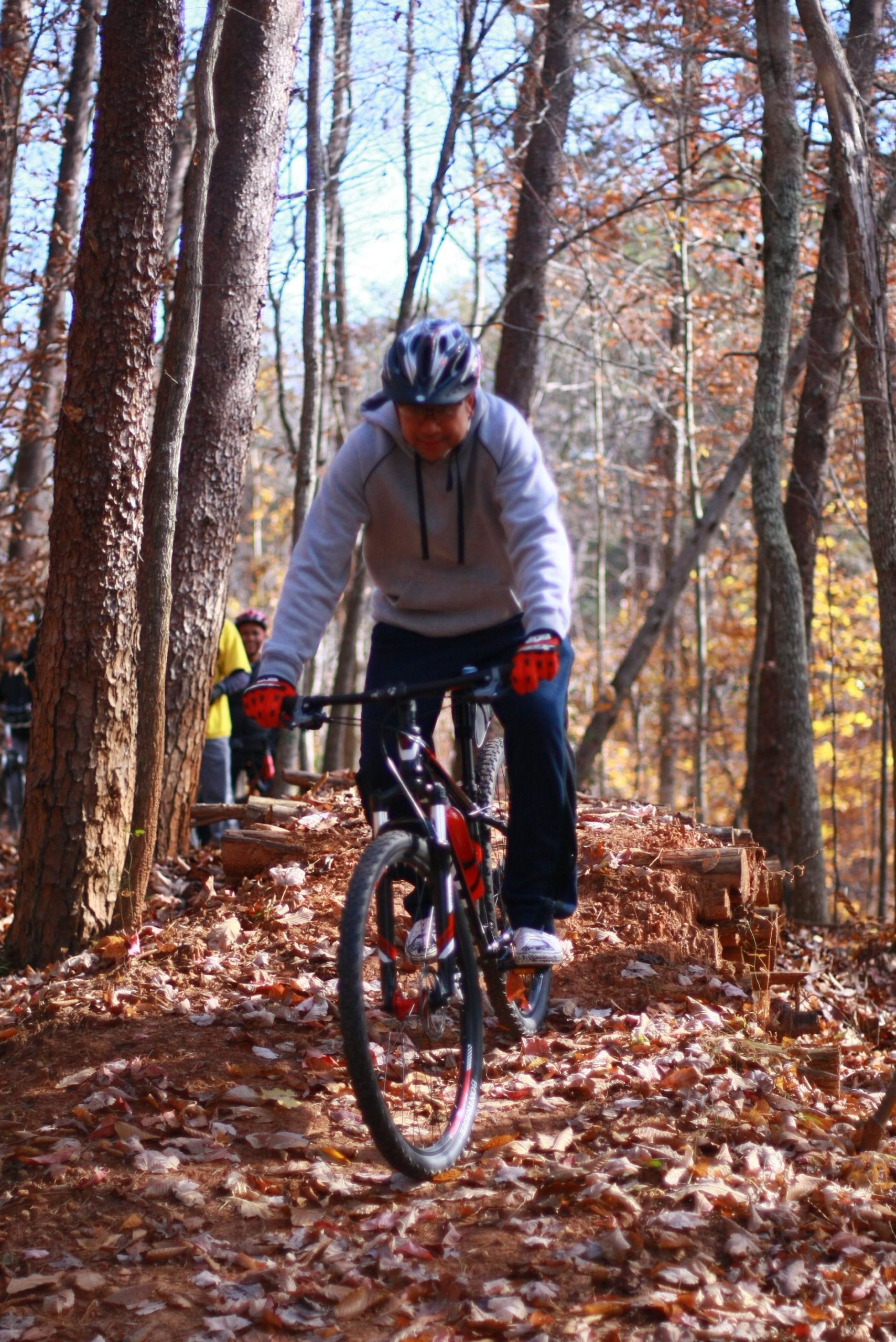 A person riding a mountain bike along a wooded trail covered with fallen leaves, surrounded by trees in autumn. The rider is wearing a helmet and a grey hoodie, leaning forward on the handlebars as they navigate the path. In the background, other cyclists can be seen. Cedar Ridge Chatmoss mountain bike trail.