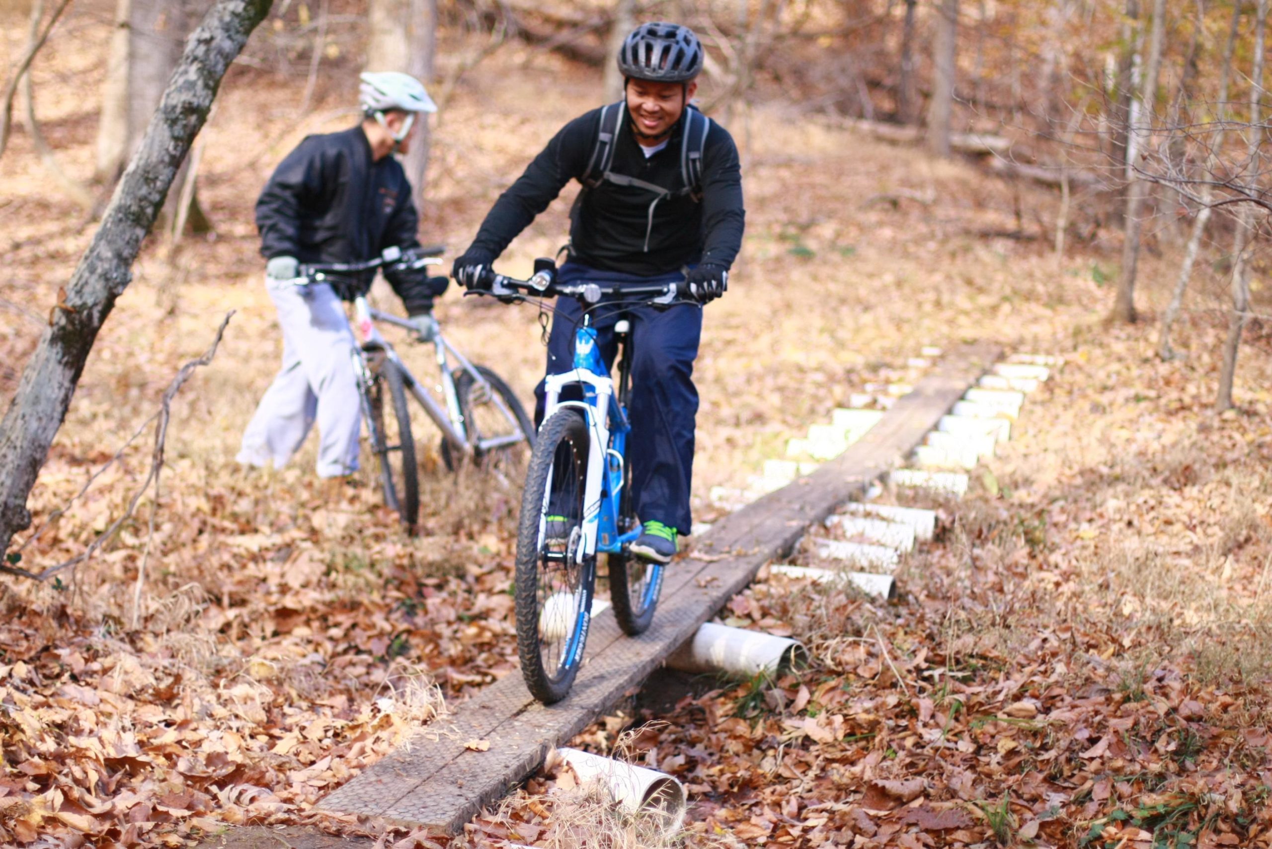 A person riding a blue mountain bike on a wooden path surrounded by autumn foliage. Another cyclist stands beside their bike, dressed in a black jacket and gray pants, observing the rider. The scene is set in a natural outdoor environment with fallen leaves covering the ground. Cedar Ridge Chatmoss mountain bike trail.