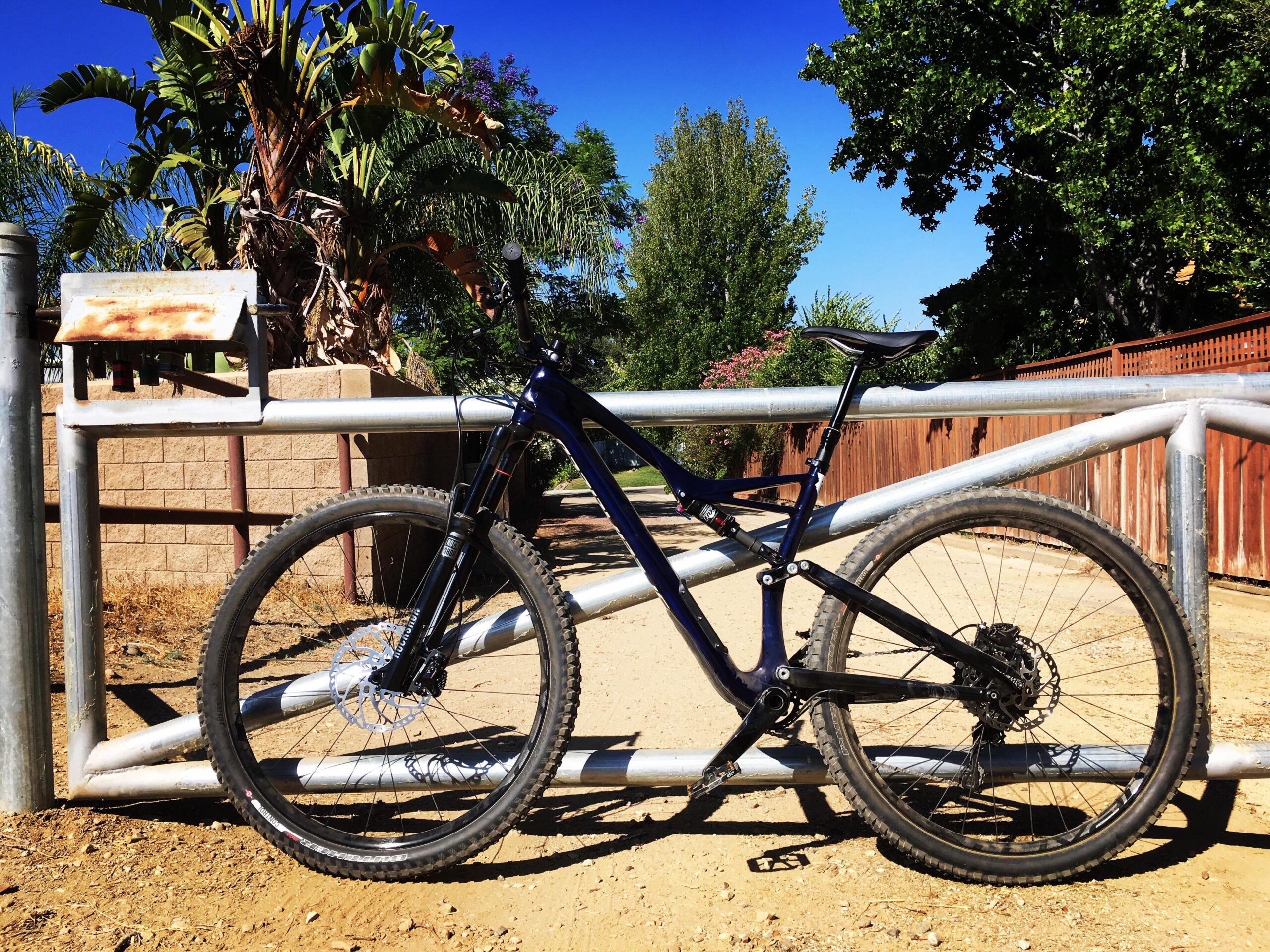Specialized Stumpjumper: A mountain bike leaning against a metal gate in a dirt pathway, surrounded by lush greenery and trees under a clear blue sky.