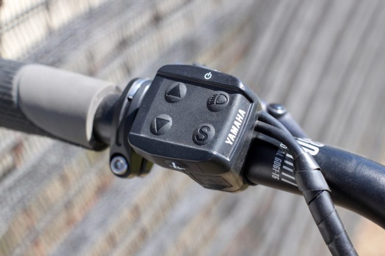 Close-up of a Yamaha control panel on a bicycle handlebar, featuring buttons for power, speed adjustment, and a selector switch, with textured grips and cables visible.