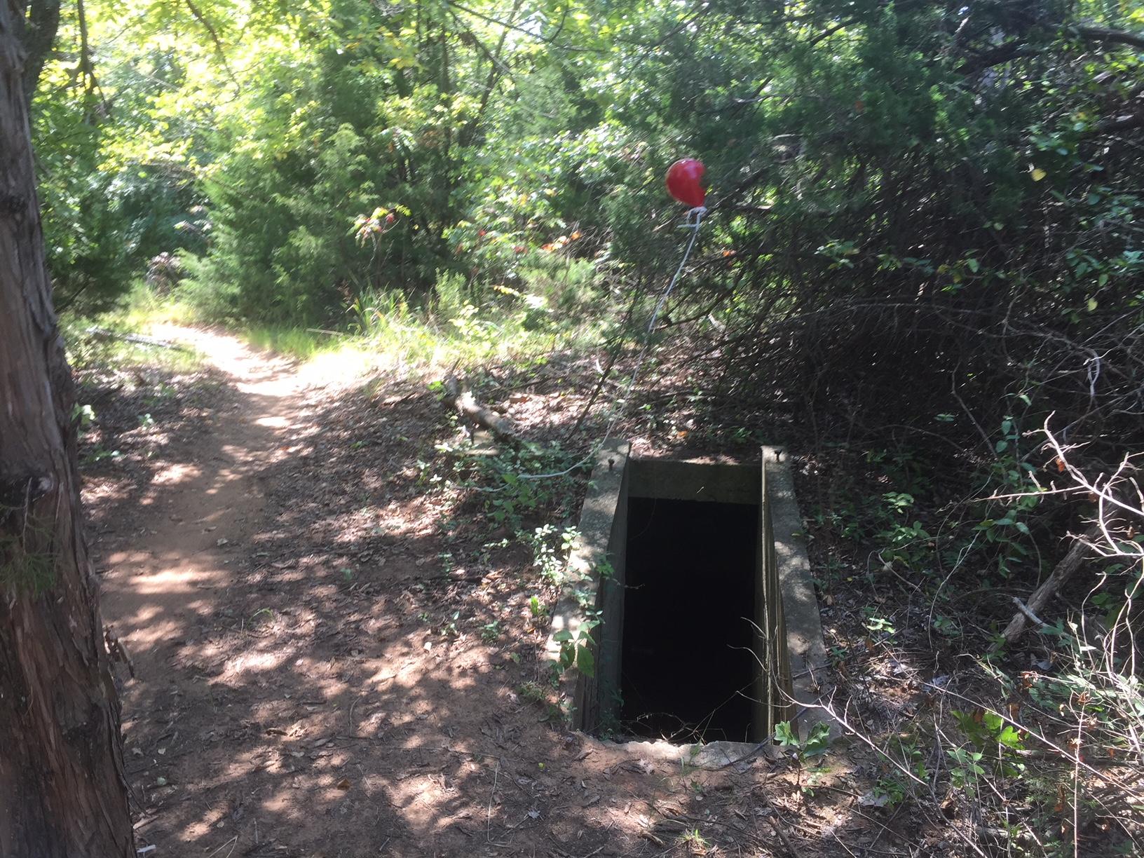 A secluded dirt path surrounded by dense greenery leads to a dark, rectangular opening, likely an abandoned well or drainage. A red balloon is tethered nearby, adding a contrasting pop of color against the natural setting. Lake Stanley Draper mountain bike trail.