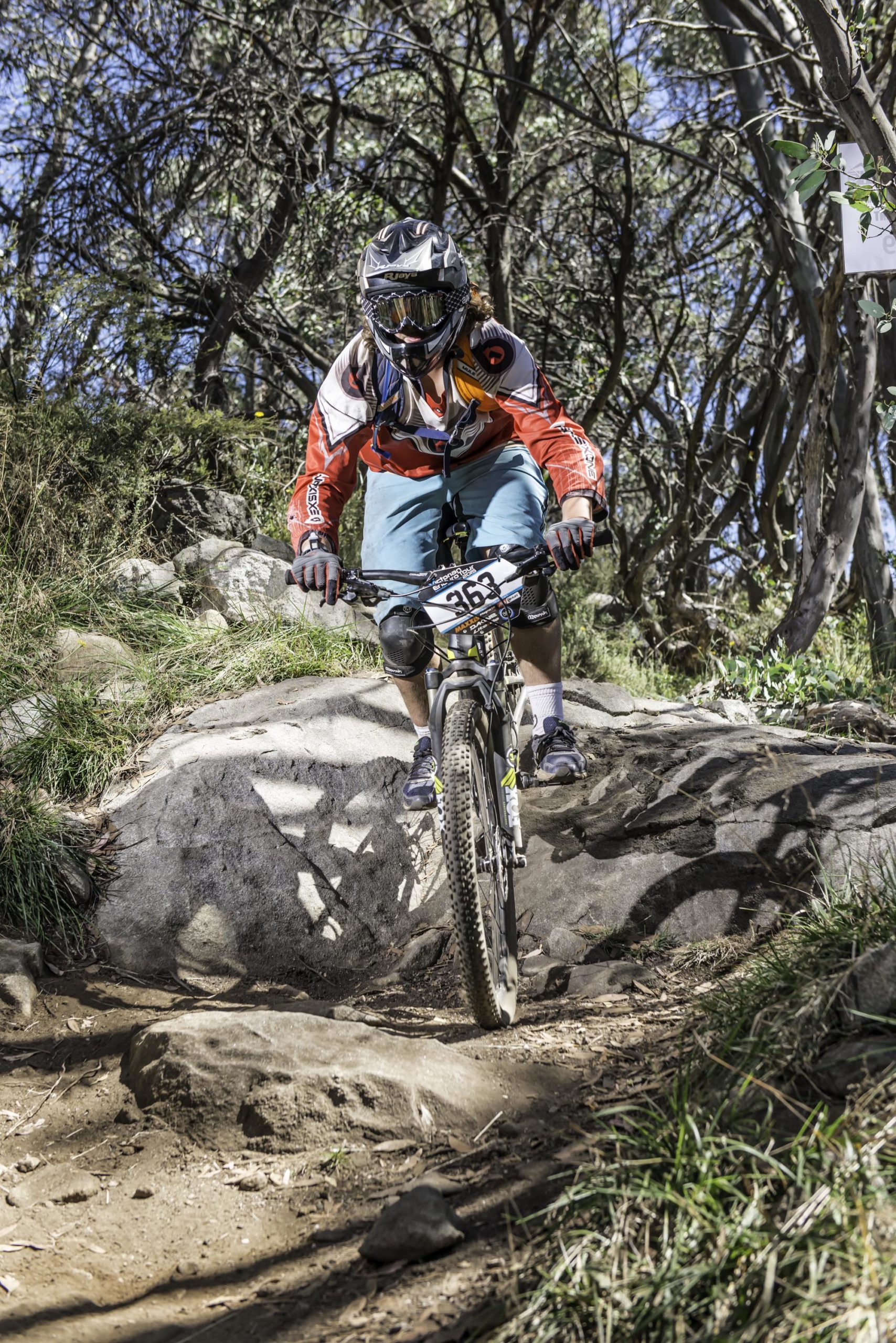 A mountain biker navigating a rocky trail, wearing a helmet, goggles, and protective gear. The rider is in a dynamic pose with their bike positioned over a large rock, surrounded by greenery and trees under a clear blue sky. Vortex mountain bike trail.