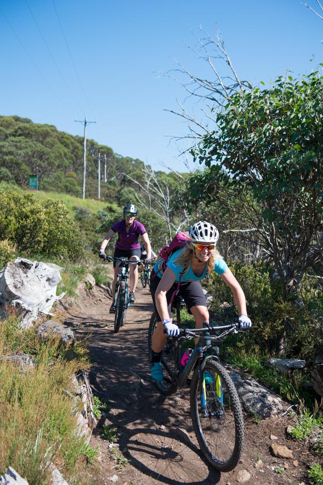 Two mountain bikers navigating a rocky trail through a lush green landscape under a clear blue sky. The first biker, wearing a helmet and colorful gear, is smiling as they ride, while the second biker follows closely behind. The scene conveys a sense of adventure and outdoor activity. Induction mountain bike trail.