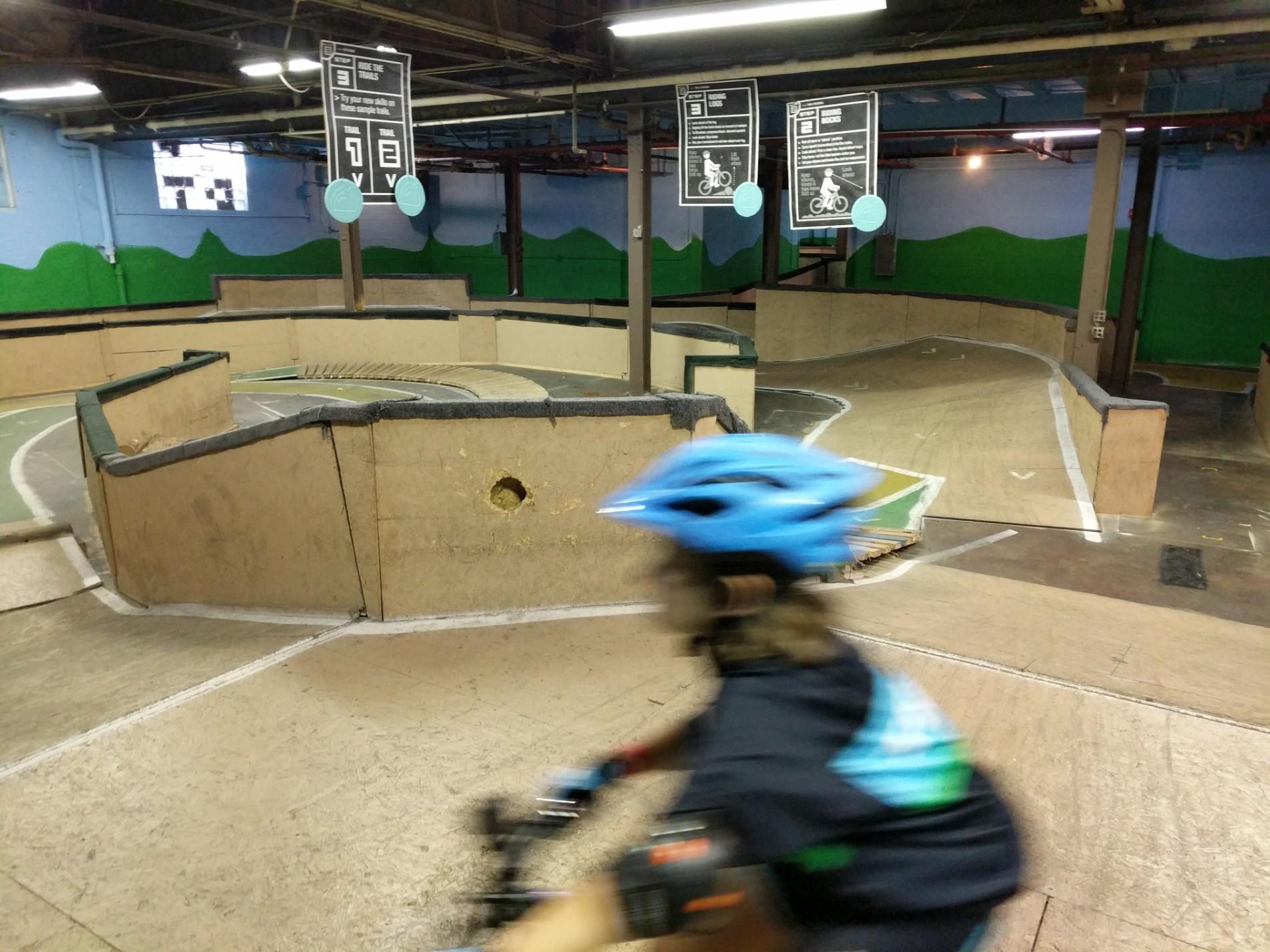 A child wearing a blue helmet rides a bicycle on an indoor BMX track. The track features wooden barriers, slopes, and circular turns. In the background, there are signs displaying scores and rules. The walls are painted with green hills, and overhead lights illuminate the space. The child is captured in motion, suggesting a dynamic riding experience. Rays Indoor Mtb Park mountain bike trail.