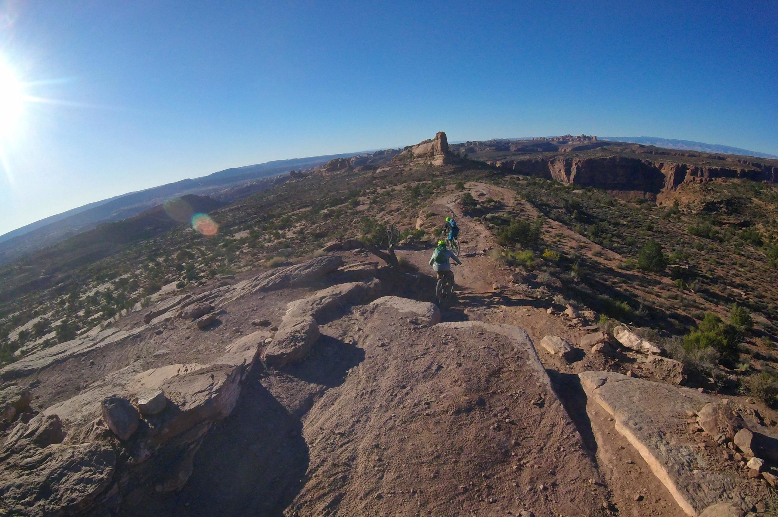 Two mountain bikers riding on a rocky trail along a canyon rim under a clear blue sky, with the sun shining brightly in the upper left corner. The landscape features scattered shrubs and distant rock formations. Porcupine Rim mountain bike trail.