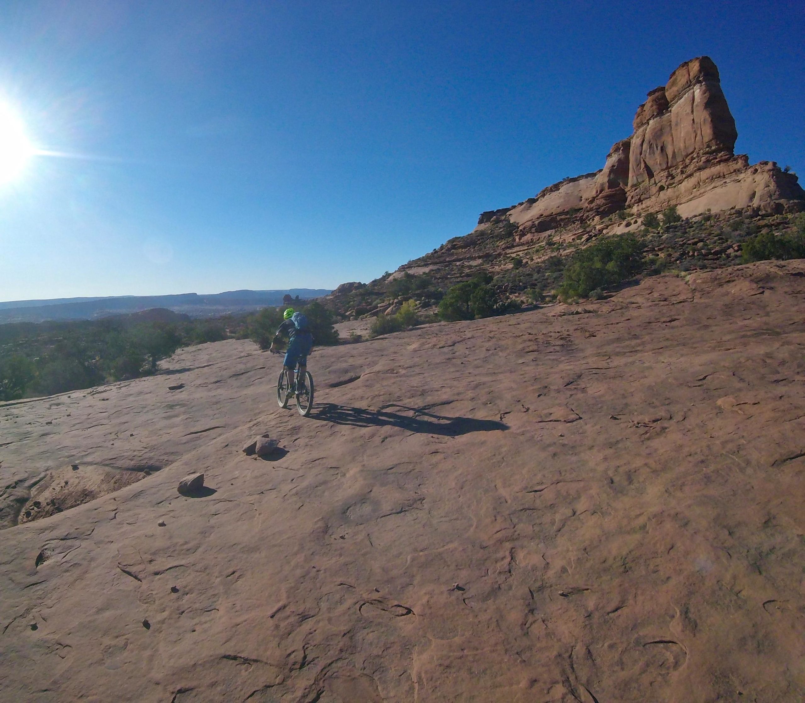 A mountain biker riding on a rocky desert landscape under a clear blue sky, with sunlight shining in the upper left corner and a prominent rock formation in the background. Porcupine Rim mountain bike trail.