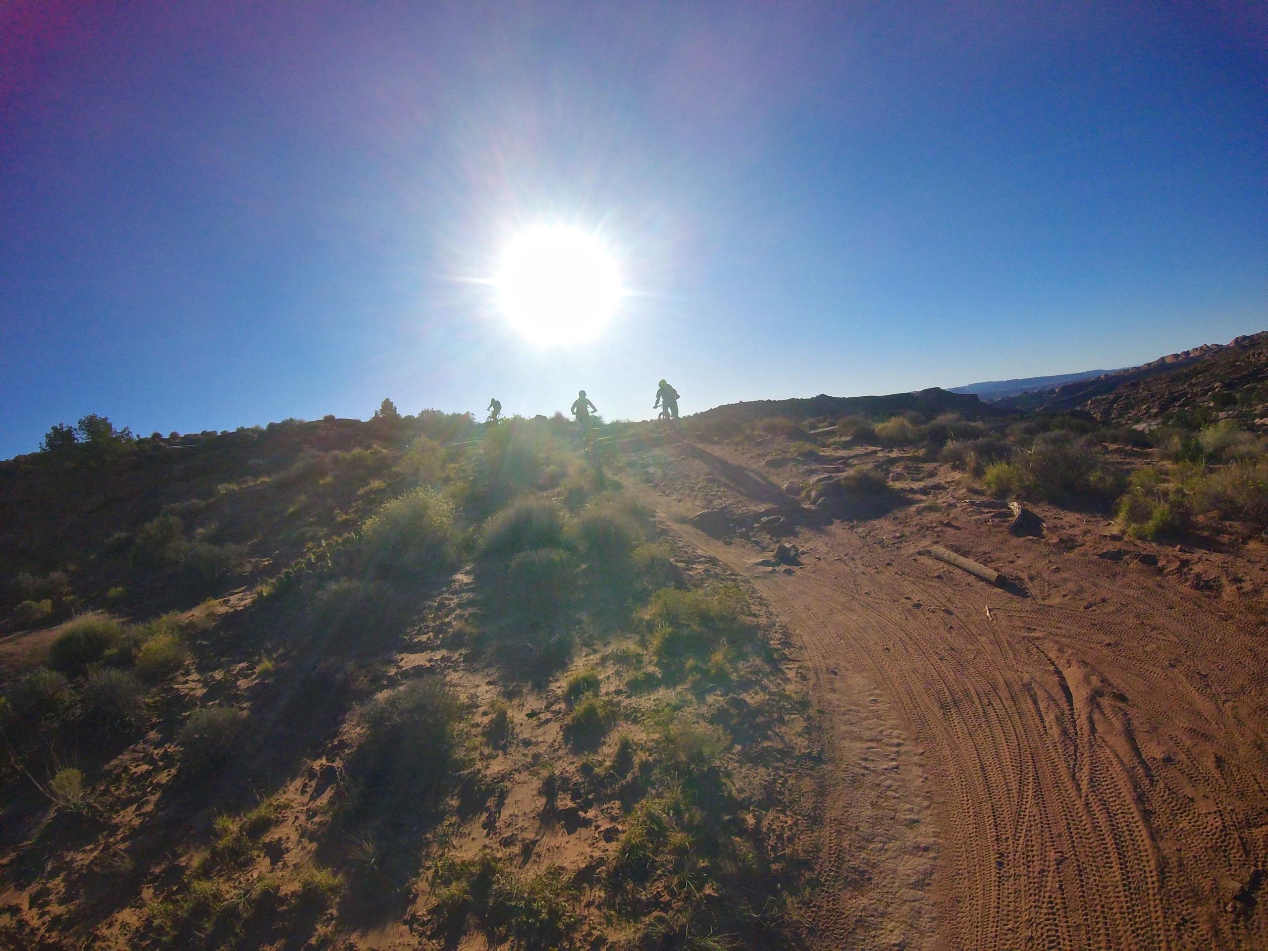 Two mountain bikers silhouetted against a bright sun on a dirt trail surrounded by sparse vegetation and rocky terrain, with a clear blue sky in the background. Porcupine Rim mountain bike trail.