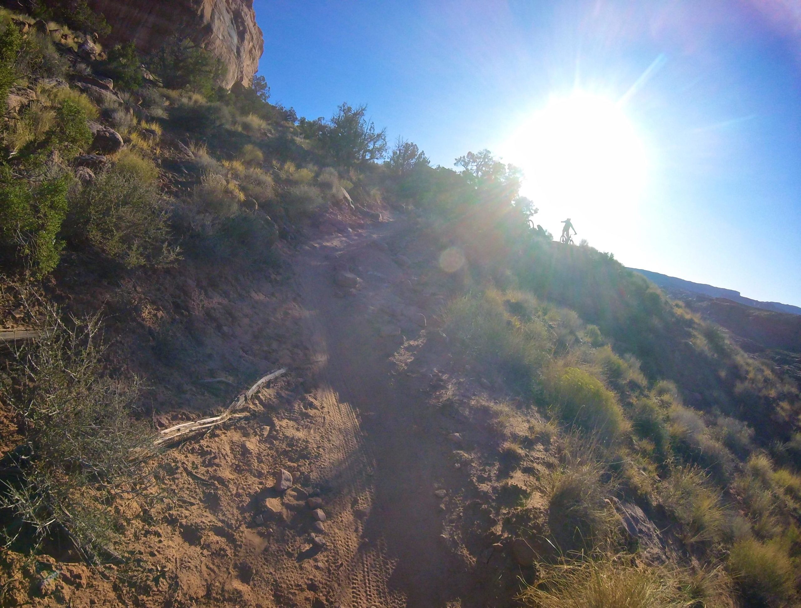 A sunlit hiking trail winding up a rocky slope, surrounded by shrubs and grasses. In the distance, a person is visible standing on a ridge, silhouetted against the bright sun. The scene captures the beauty of nature and outdoor adventure. Porcupine Rim mountain bike trail.